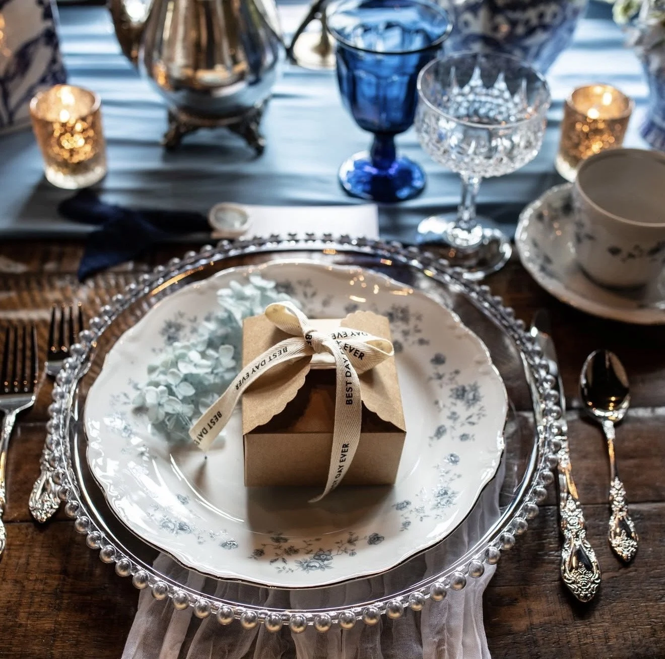 Elegant table setting with a white plate and a gift box wrapped with a ribbon on a fancy charger plate, surrounded by silverware, glassware, candles, and decorative items.