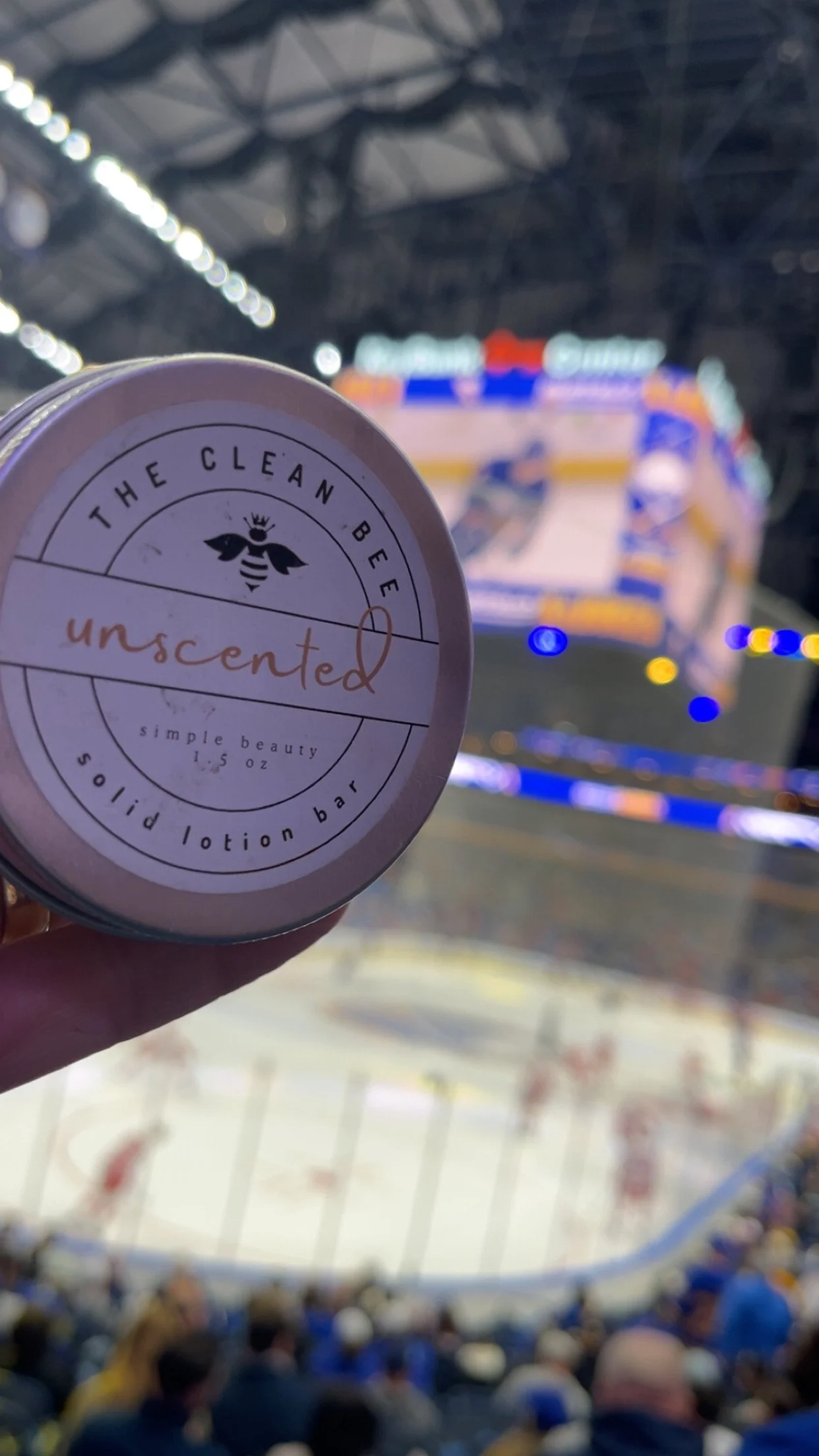 A hand holding a small round container of The Clean Bee unscented solid lotion bar in front of an ice hockey arena filled with spectators.