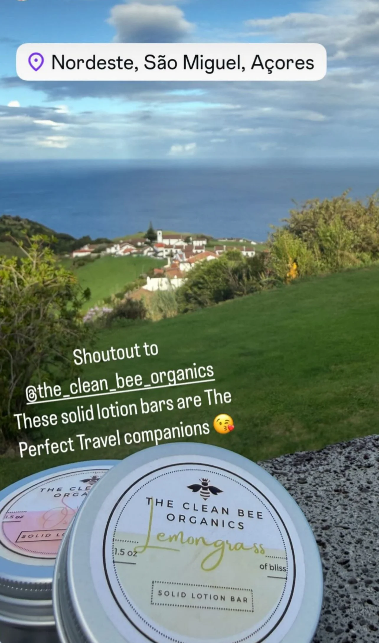 A scenic view of a small village with white buildings and red rooftops, green hills, and the ocean in the background sky partly cloudy. In the foreground, two solid lotion bars from The Clean Bee Organics, labeled Lemon Grass, are placed on a stone s