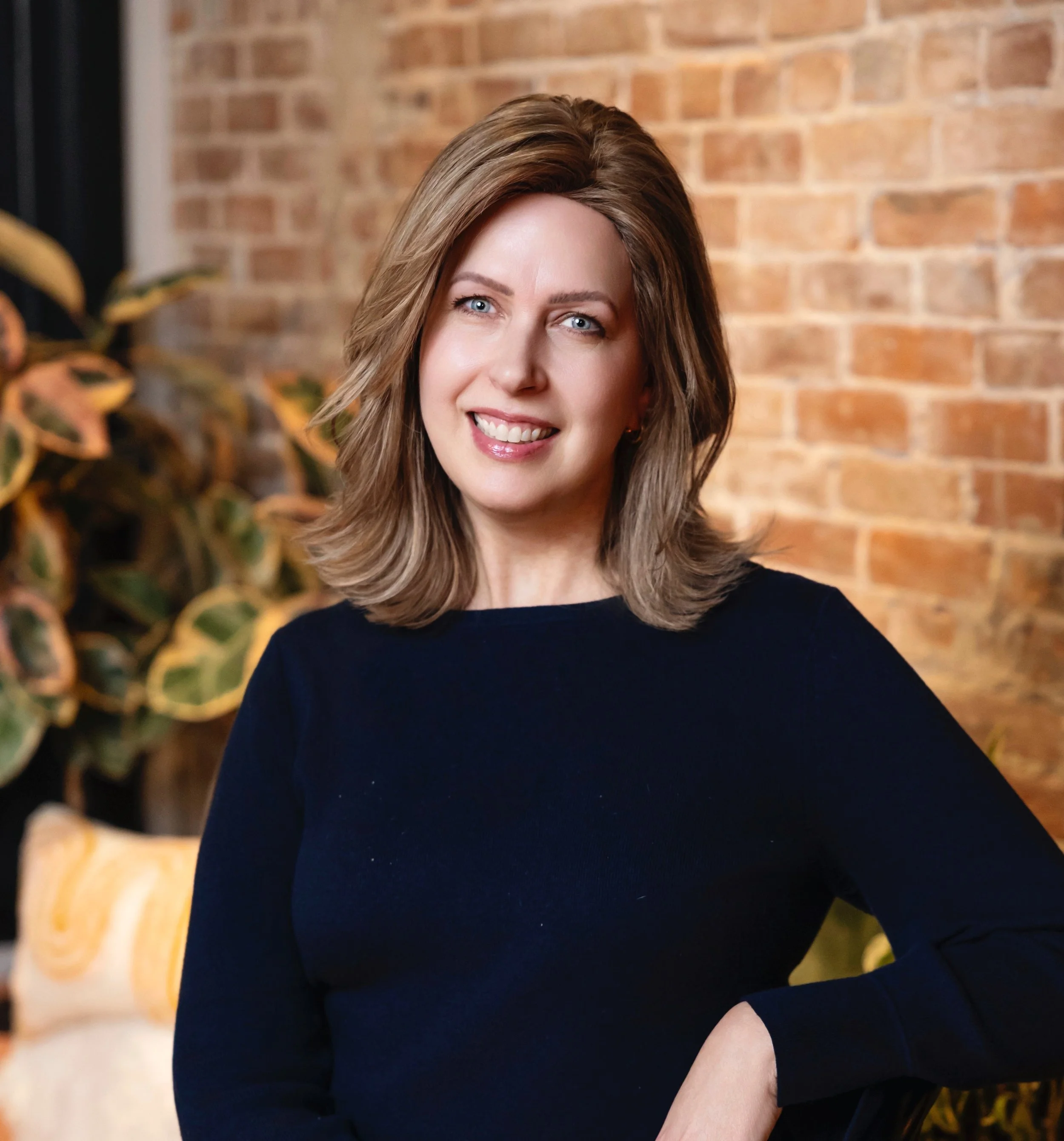 A woman with shoulder-length light brown hair, blue eyes, and a friendly smile posing indoors against a brick wall with a plant in the background.