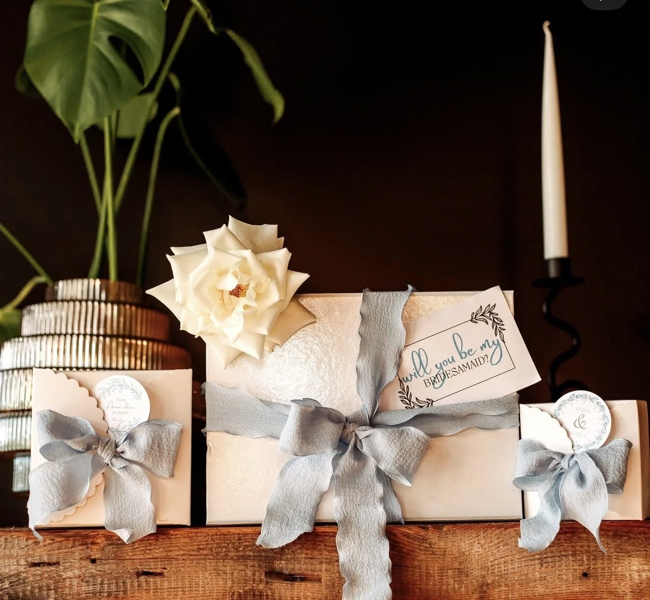 Decorative wedding or engagement gift setup with wrapped presents tied with gray ribbons, a white flower, a handwritten card asking "will you be my BRIDE SAMAID?", a potted plant with large green leaves, and a lit white candle in a black holder on a wooden surface.