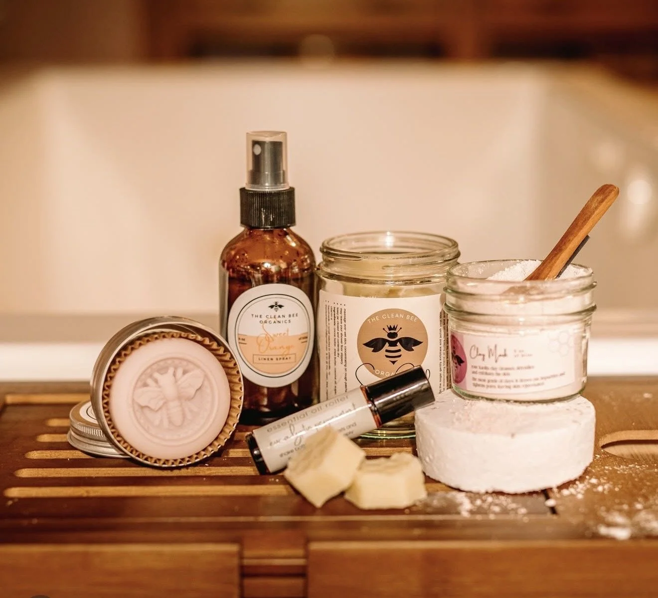 Natural skincare products on a wooden surface, including a spray bottle, a jar of loose powder with a wooden spoon, a soap with bee design, and small cubes of soap or lotion
