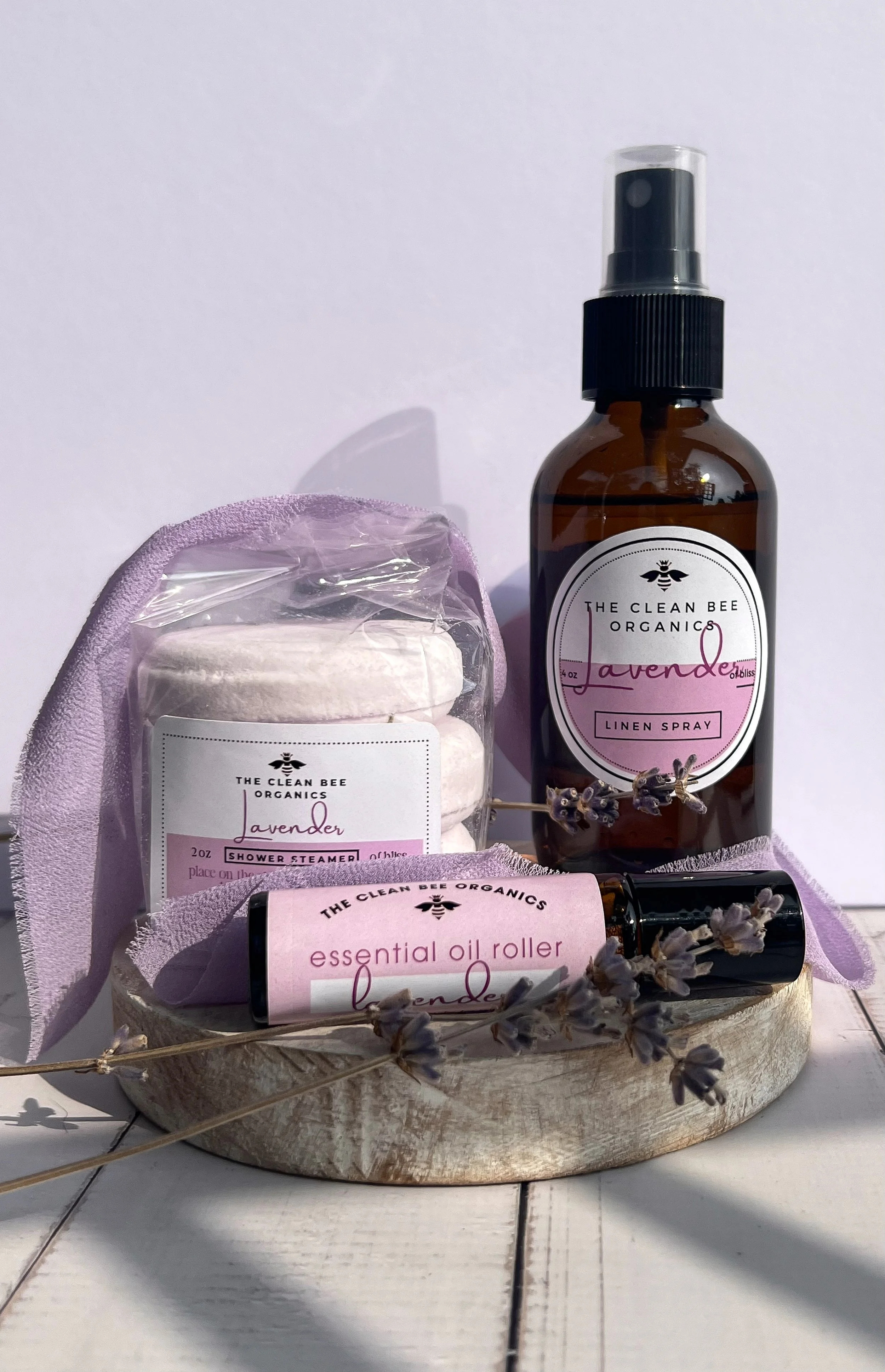 Set of lavender-scented organic personal care products including a linen spray, essential oil roller, and shower steamers on a wooden platter, decorated with sprigs of lavender and pink fabric.