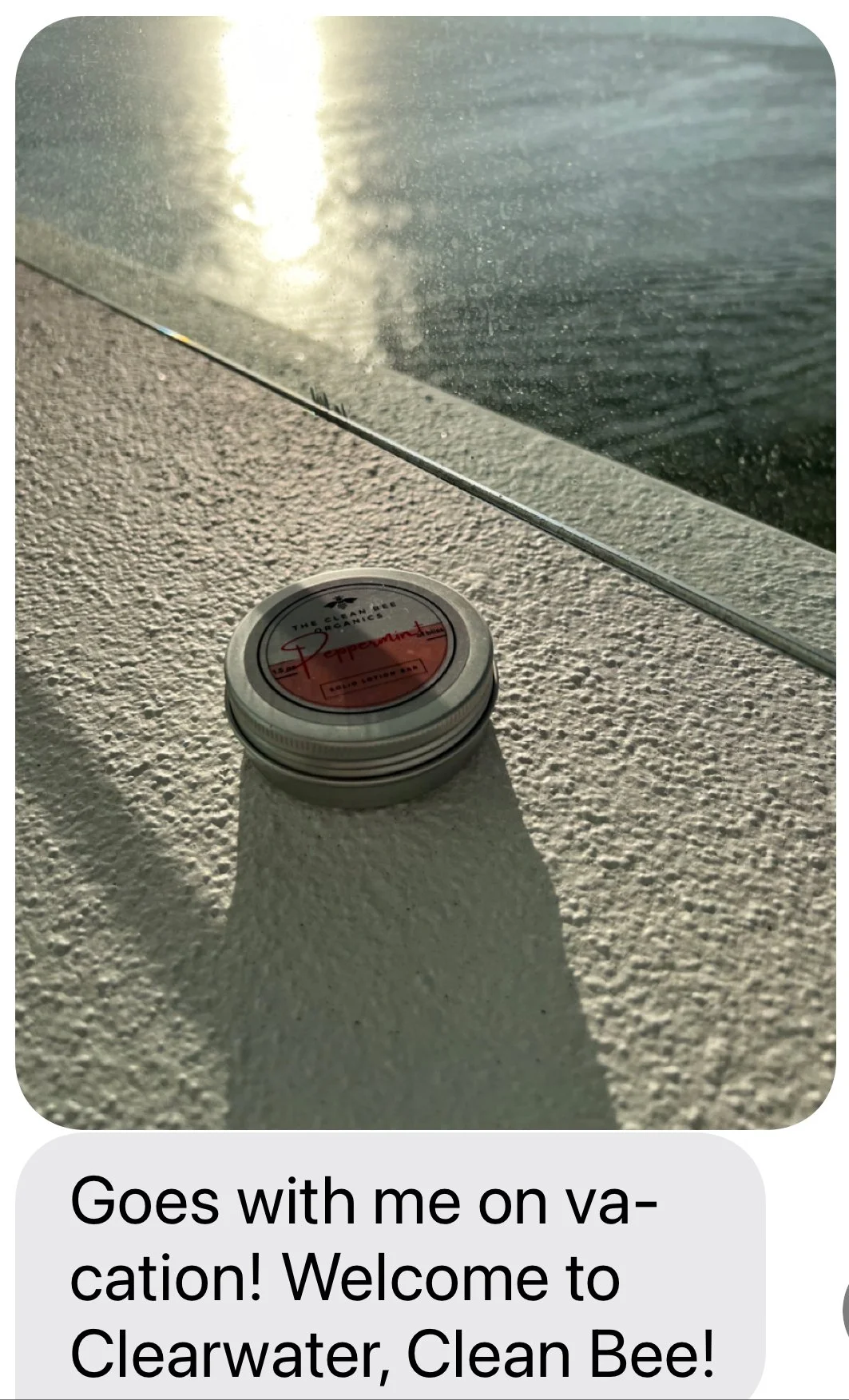 A small round tin container labeled 'The Clean Bee Organics' with a red label that says 'Peppermint' sitting on a textured white surface, with sunlight reflecting on a body of water in the background.
