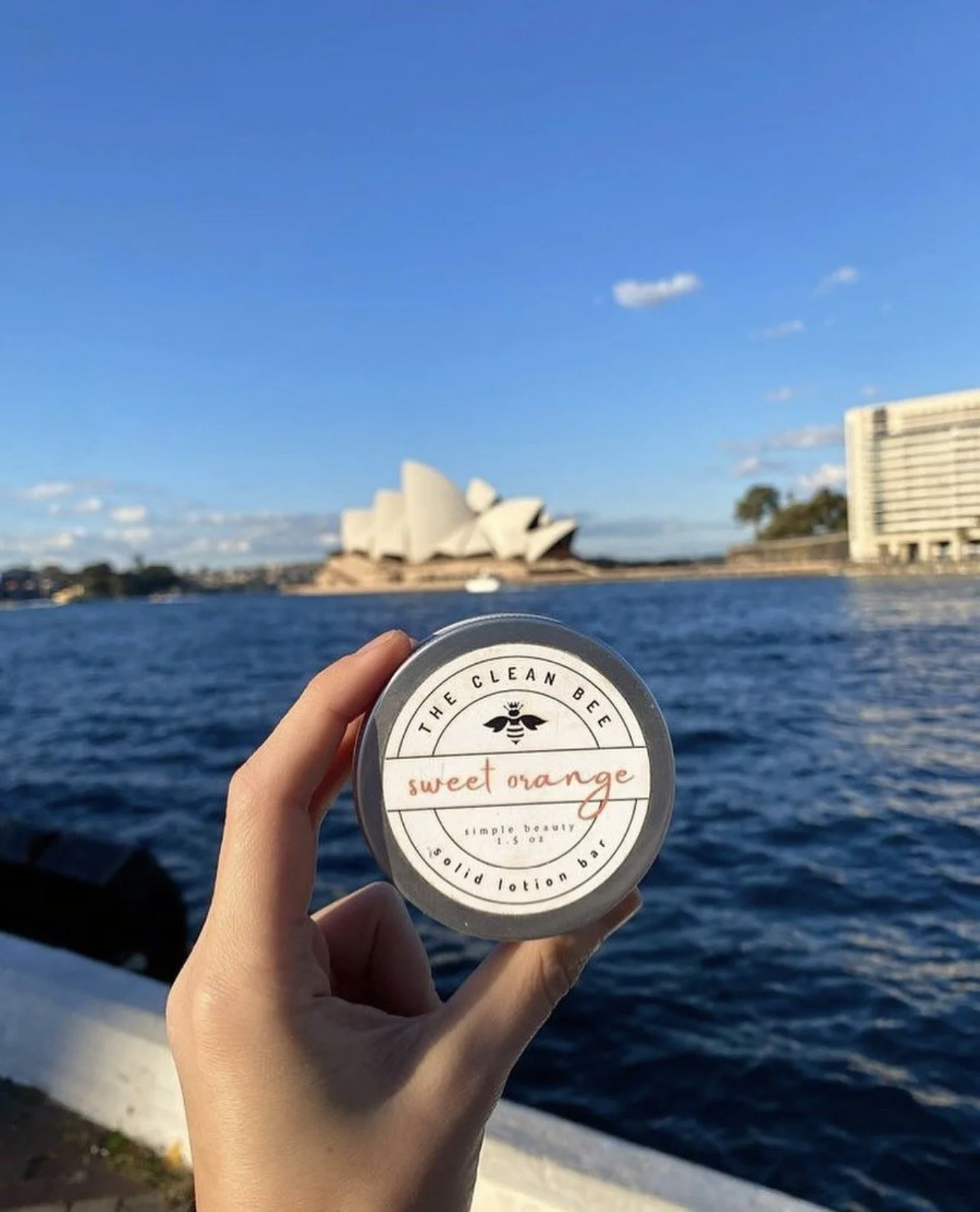 Hand holding a container of 'Sweet Orange' lotion with Sydney Opera House and Sydney Harbour Bridge in the background on a sunny day.