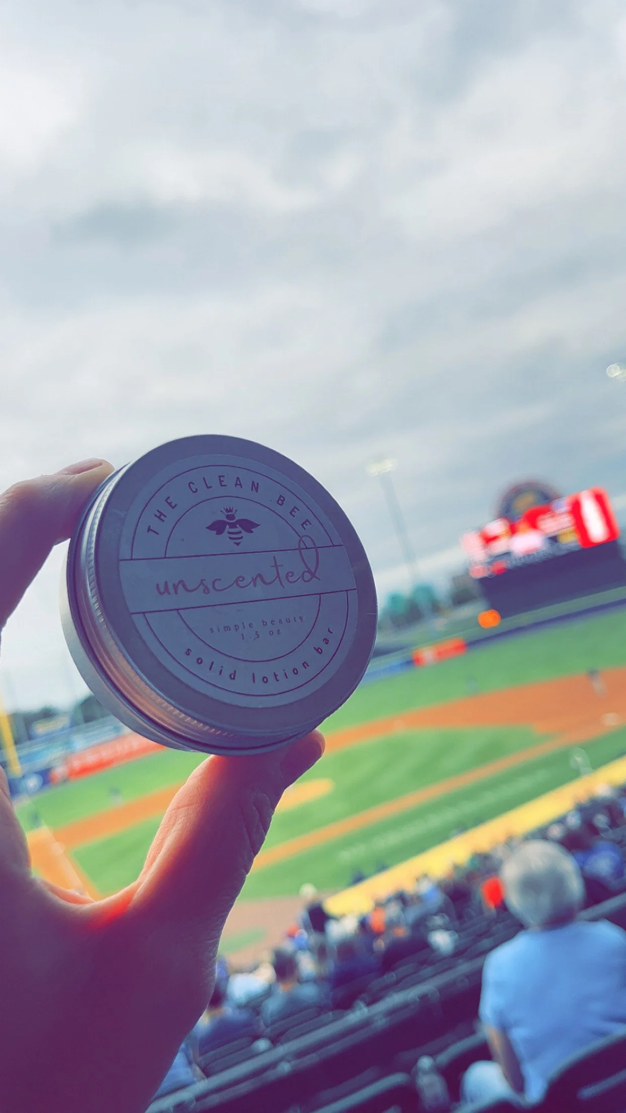 Person holding a container of unscented lotion at a baseball game, with stadium and field in the background.