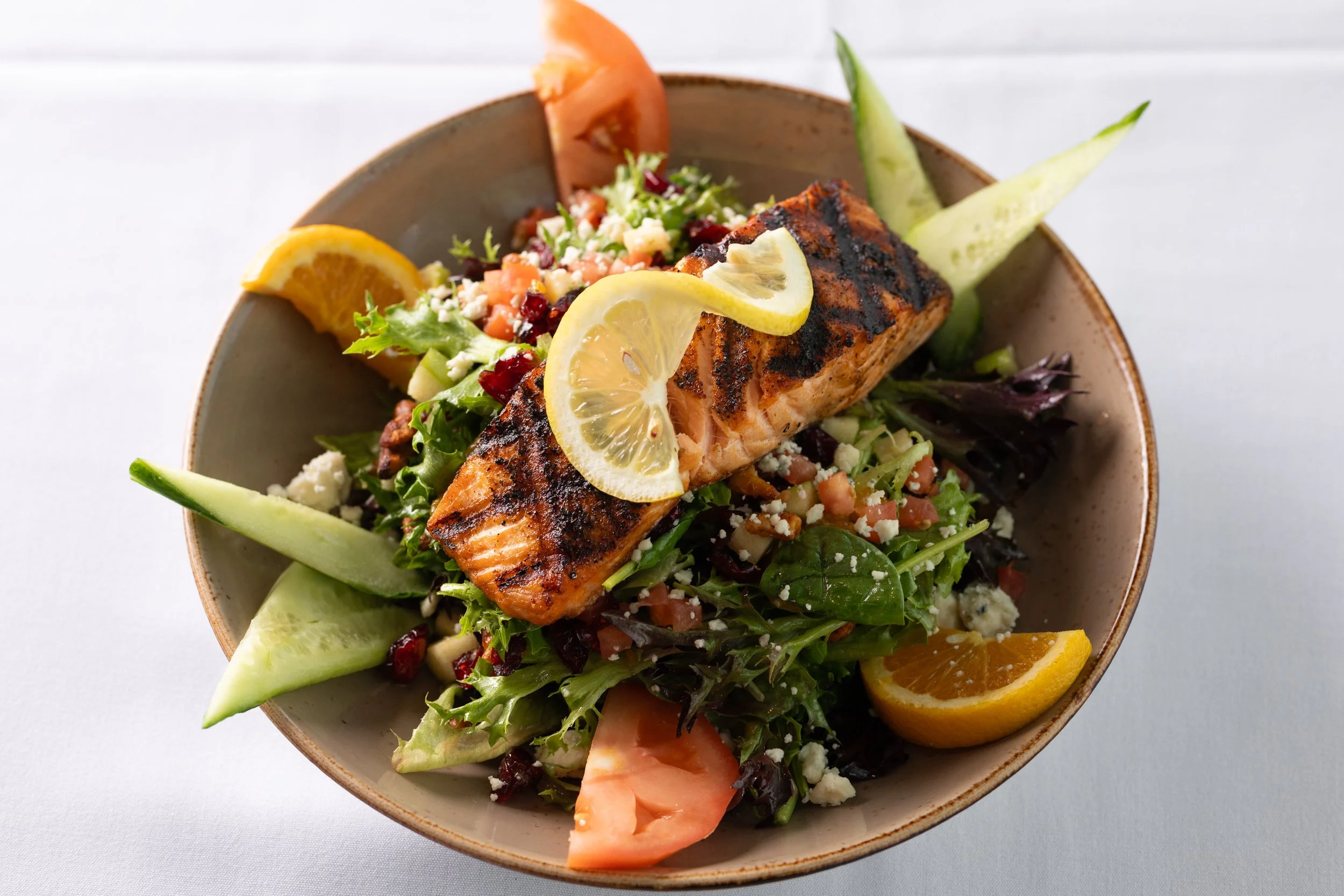 Grilled salmon on a bed of mixed greens, cucumber, tomato, pomegranate seeds, crumbled cheese, and lemon wedges in a bowl.