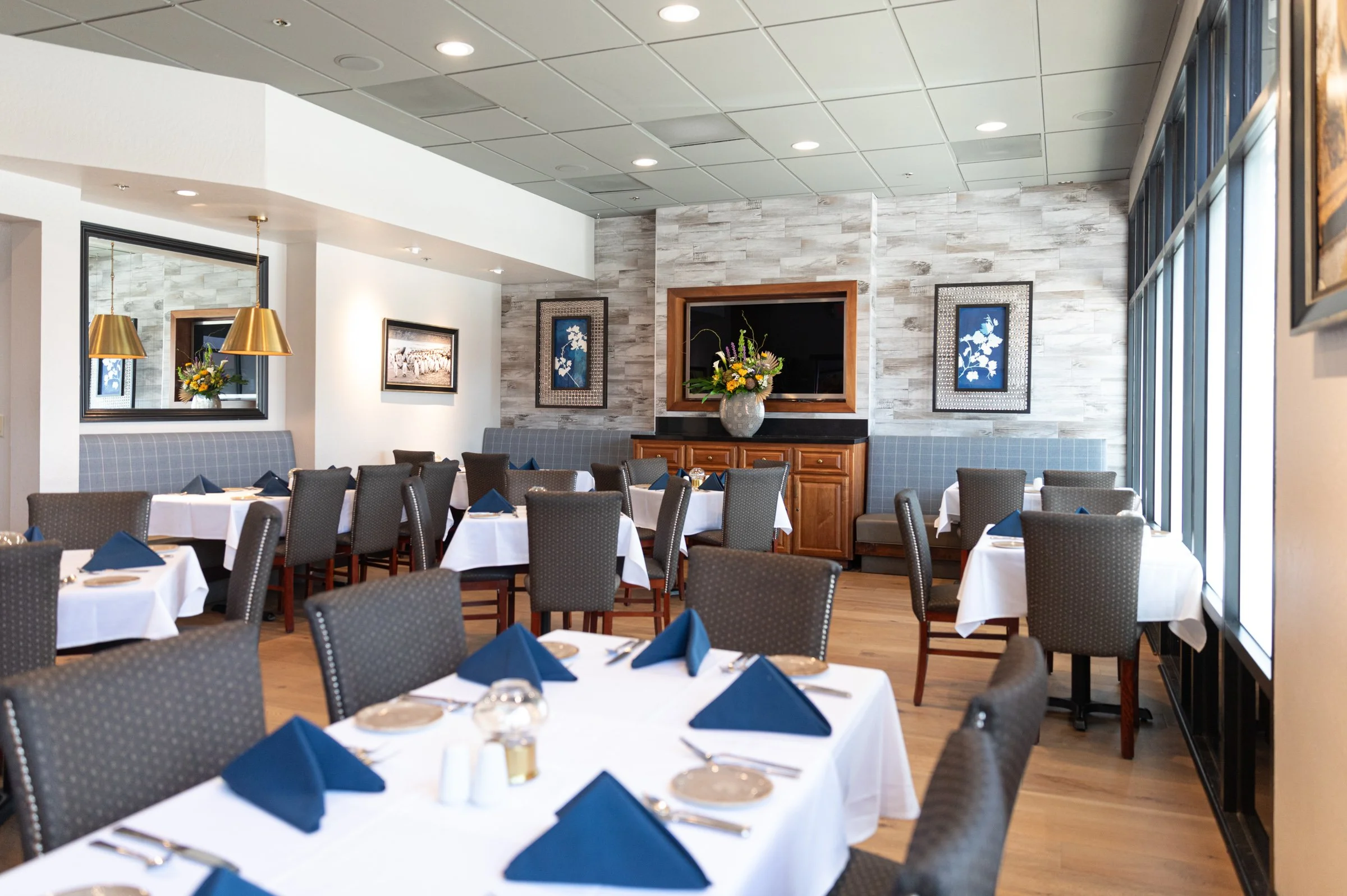 Elegant restaurant with tables set with white tablecloths, blue napkins, and silverware, featuring a gray and white color scheme, wall art, and large windows.