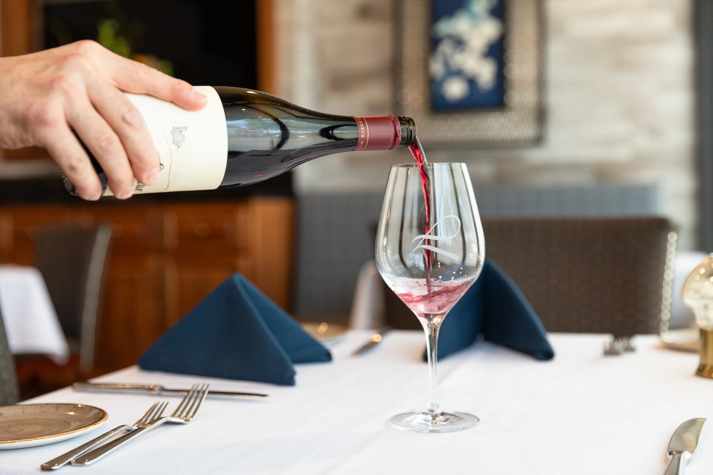 Person pouring red wine into a wine glass on a dining table with navy blue napkins, silverware, and a white tablecloth in a restaurant setting.