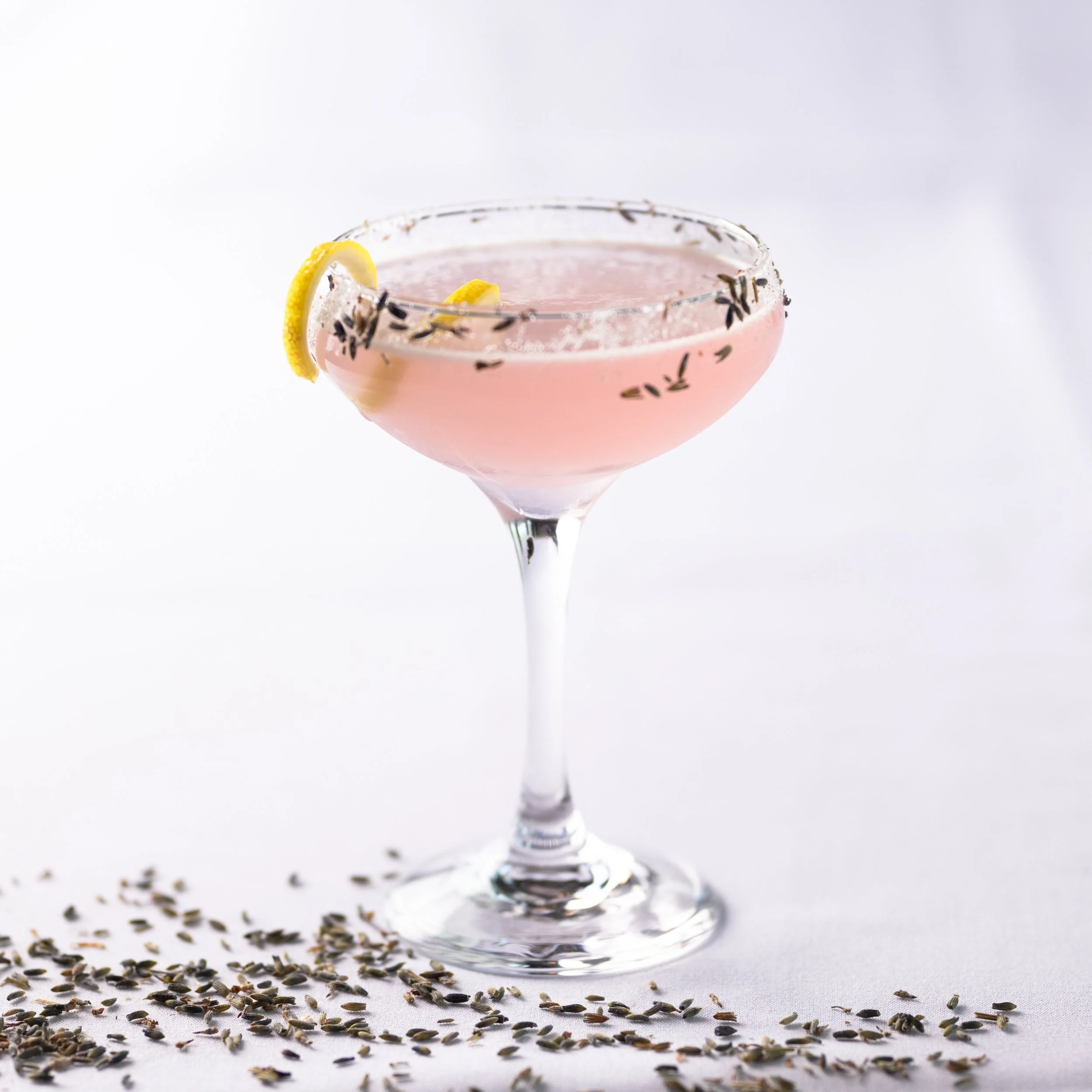A pink cocktail in a coupe glass garnished with lemon slices and a salted rim, with lavender flowers scattered around at the base on a white background.