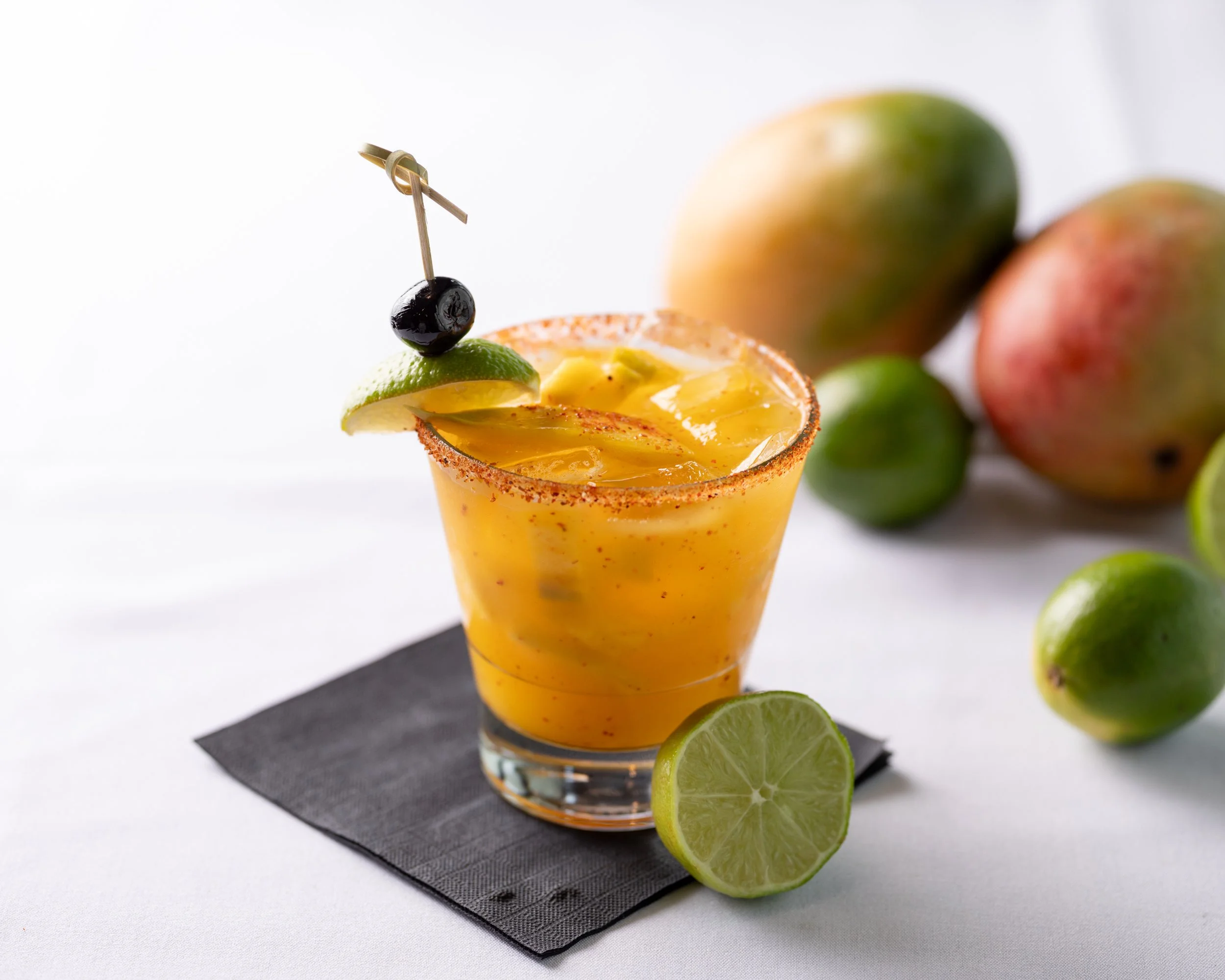 Yellow cocktail with lime wedge and black cherry garnish on a skewer, served in a salted rim glass, with whole and sliced limes and mangoes in the background.