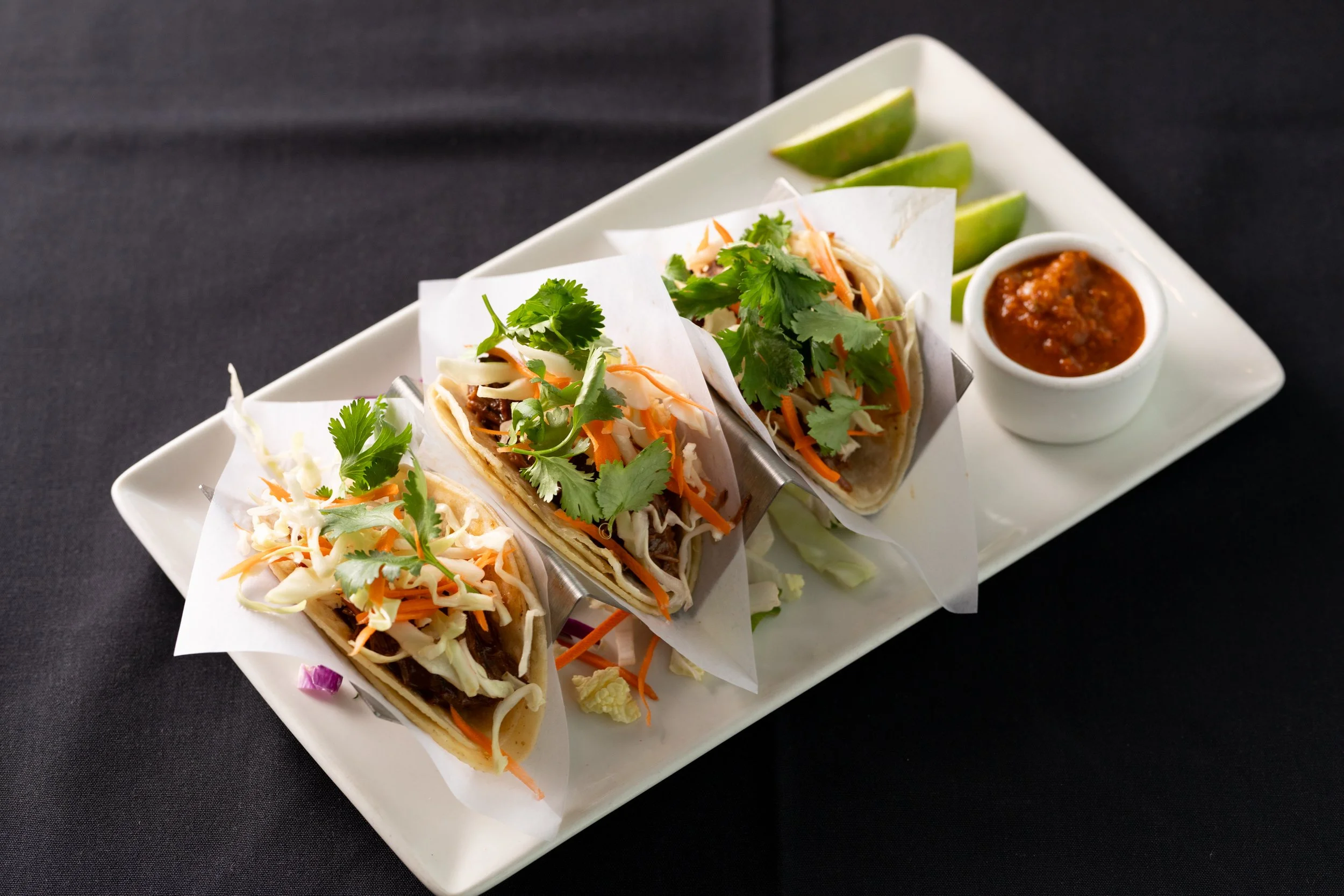 Three tacos with shredded lettuce, carrots, cilantro, and meat, served on a white rectangular plate with lime wedges and a side of salsa.