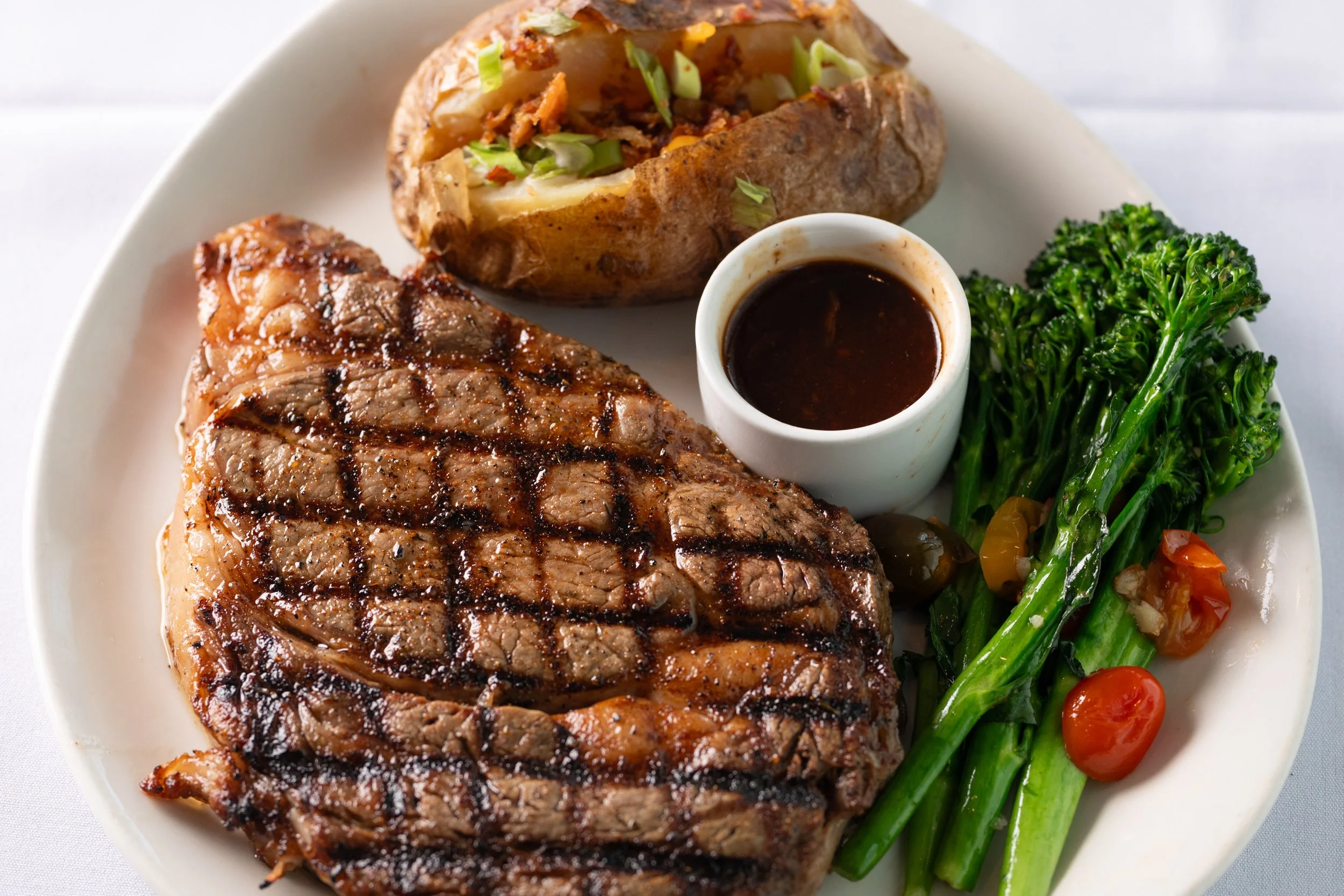Grilled steak with crosshatch grill marks, baked potato topped with green onions and bacon bits, side of steamed broccoli, cherry tomatoes, and a cup of brown gravy on a white plate.