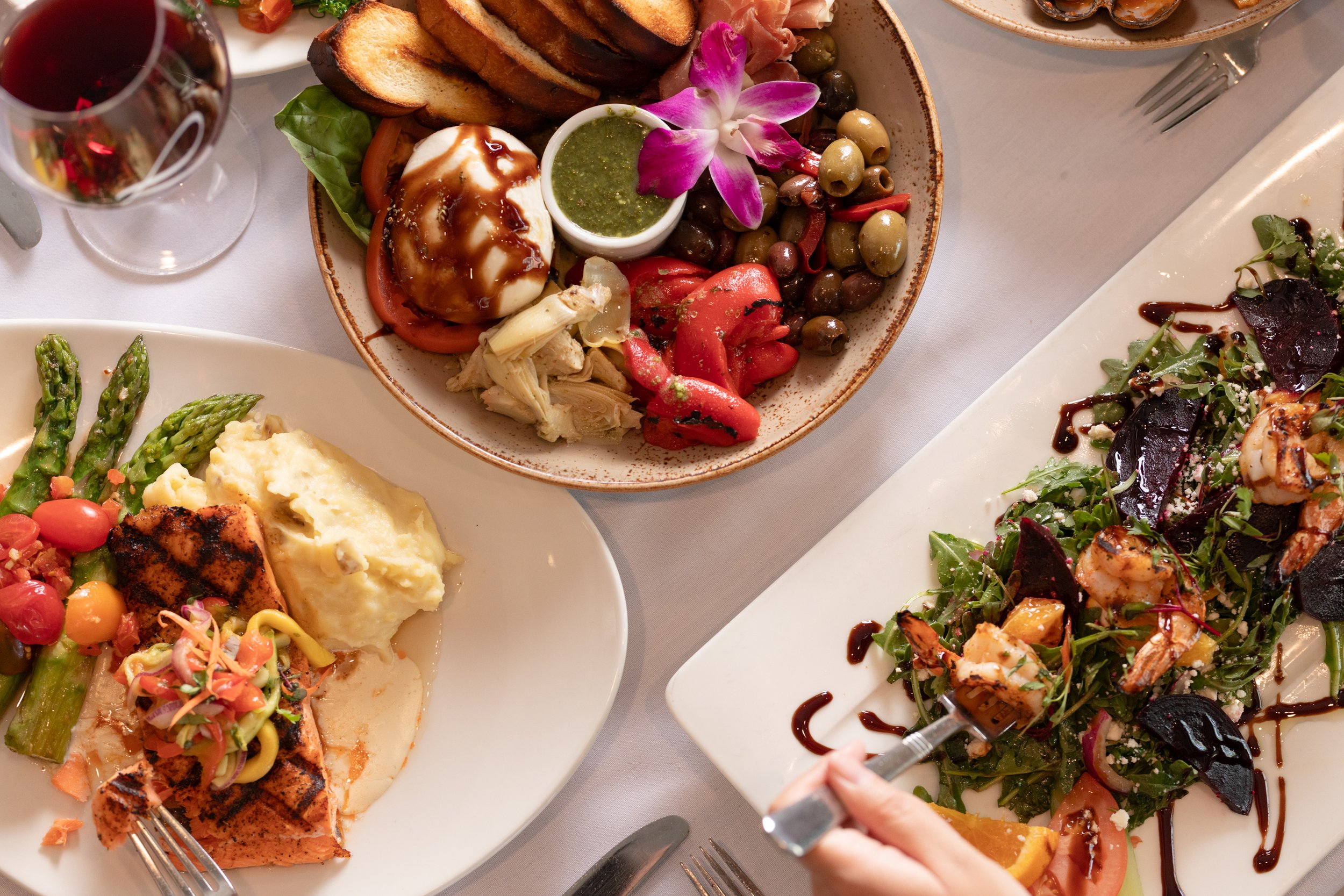 A variety of gourmet dishes on a table, including a salad with grilled shrimp and balsamic glaze, mashed potatoes with asparagus and grilled salmon, and a bowl of mixed olives, vegetables, cheese, and an edible flower.