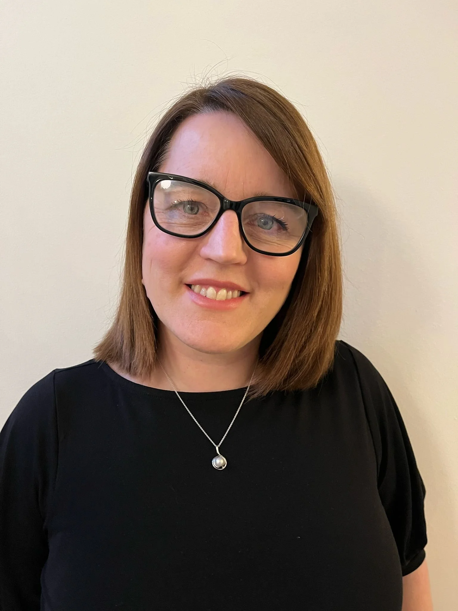 A woman with shoulder-length brown hair wearing black-rimmed glasses, a black top, and a pearl pendant necklace smiling against a plain light-colored wall.