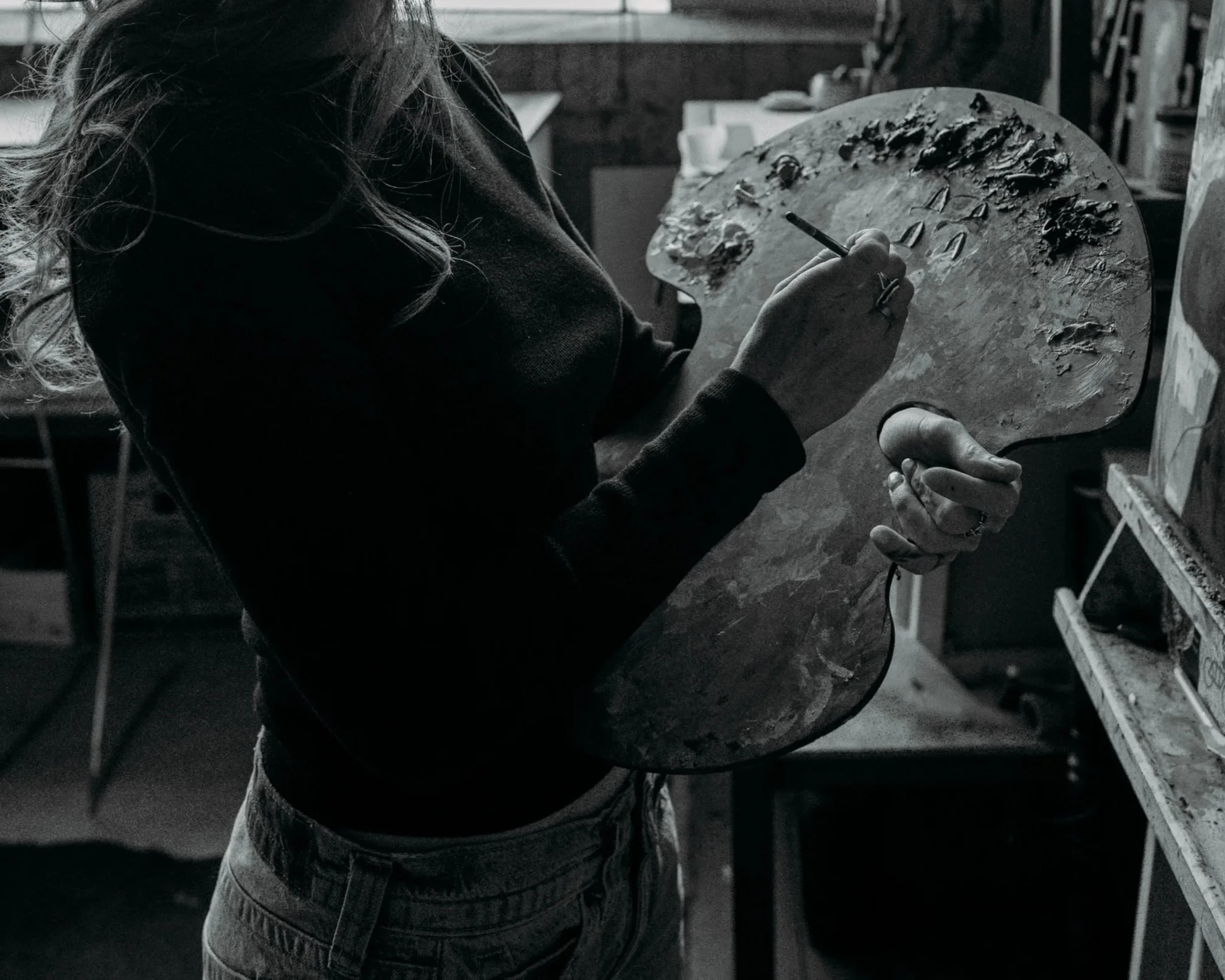 A woman painting on a palette in an art studio, holding a paintbrush and wearing a dark top and light-colored pants, surrounded by art supplies and canvases.