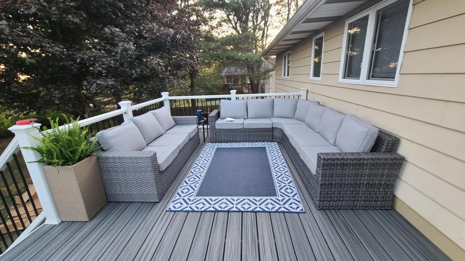 Outdoor weathered wooden deck with wicker sectional sofa with light gray cushions, a patterned outdoor rug, a potted plant, and a view of trees and neighboring house.