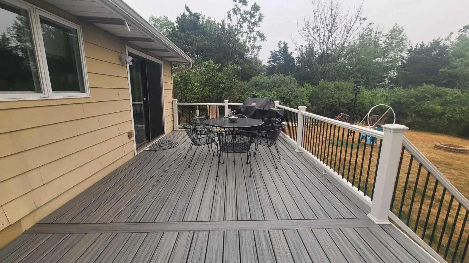 A wooden deck attached to a house with yellow siding, featuring a black metal outdoor dining table and six matching chairs, a covered barbecue grill, and surrounded by a railing overlooking a yard with trees and some outdoor equipment.
