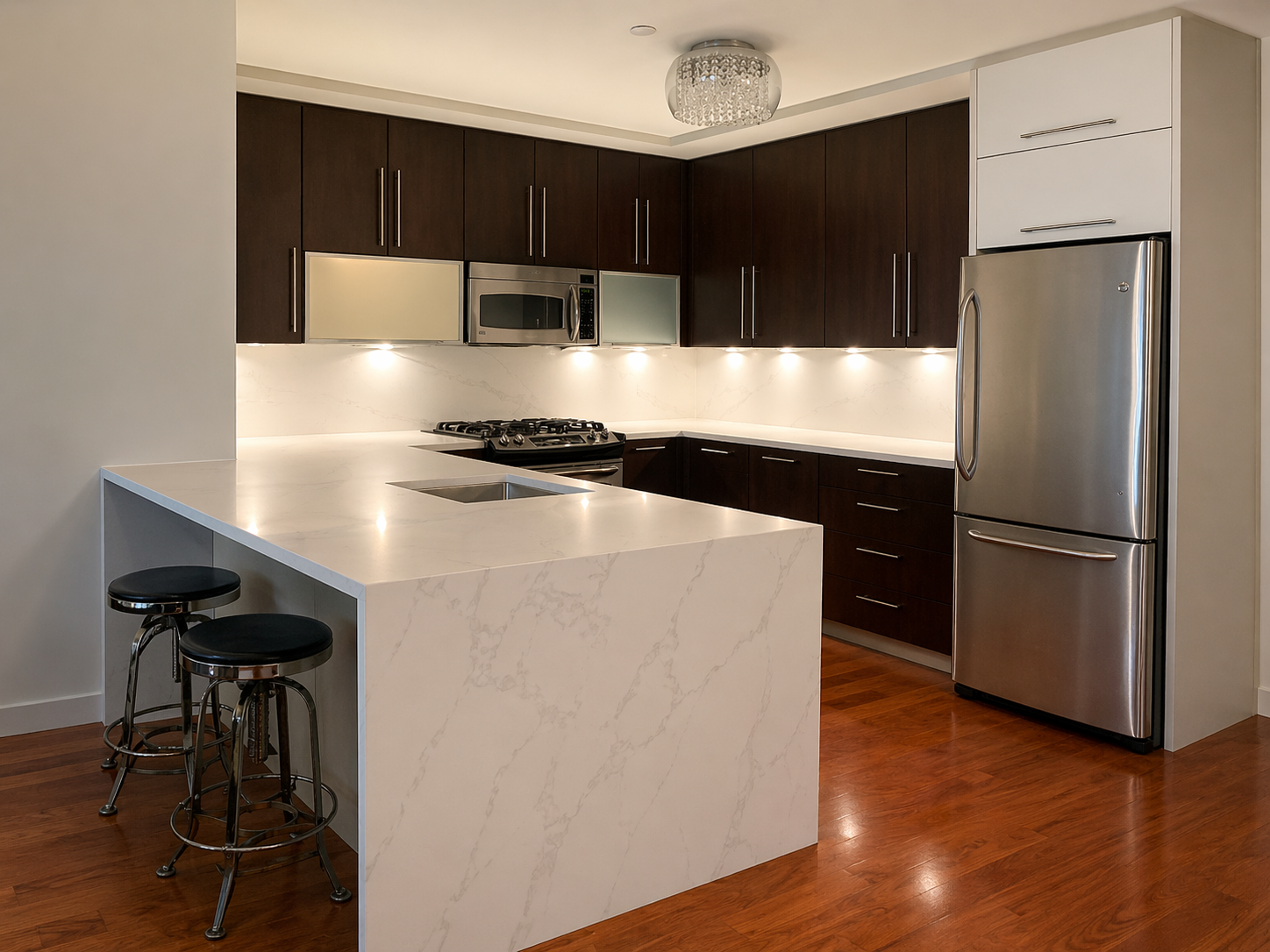 Modern kitchen with white marble island, dark wood cabinets, stainless steel refrigerator, microwave, and stove, with under-cabinet lighting and wood flooring.