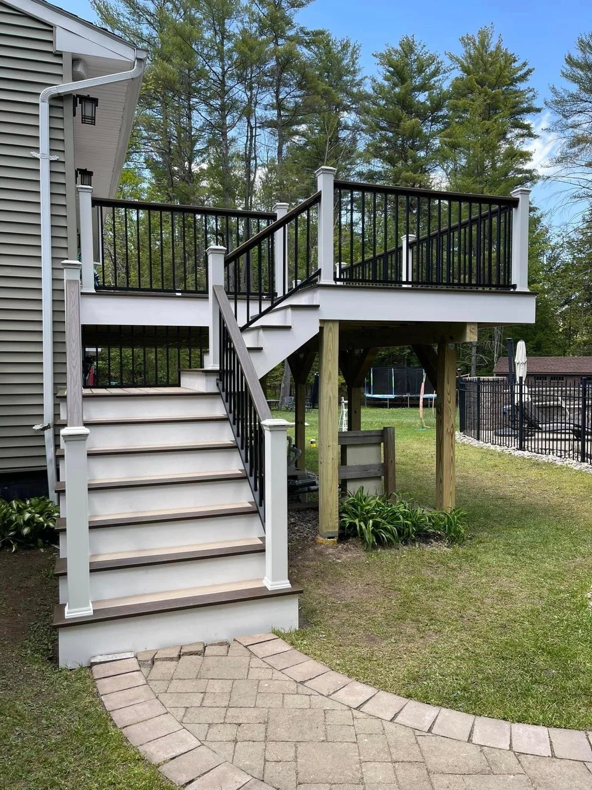 Backyard with a wooden deck and staircase. The deck has black metal railing, and a trampoline and pool are visible in the yard. There are trees and a blue sky in the background.