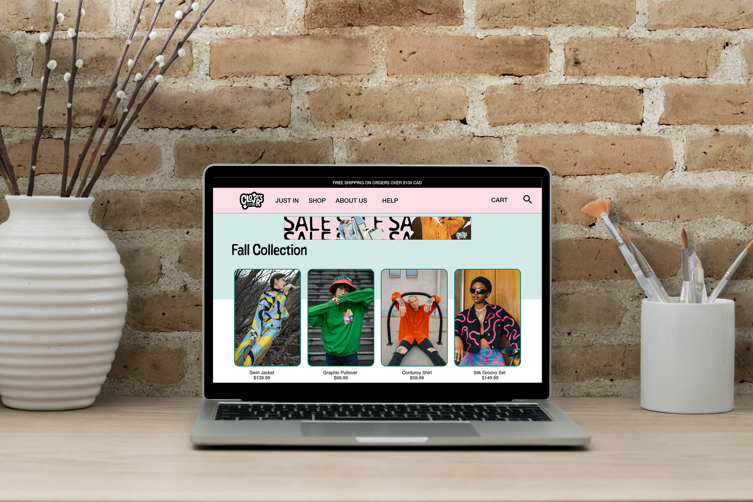 A laptop on a wooden desk displaying an online store's fall collection with images of jackets, a pullover, a shirt, and a set, against a brick wall background, flanked by white vases with decorative sticks and paintbrushes.