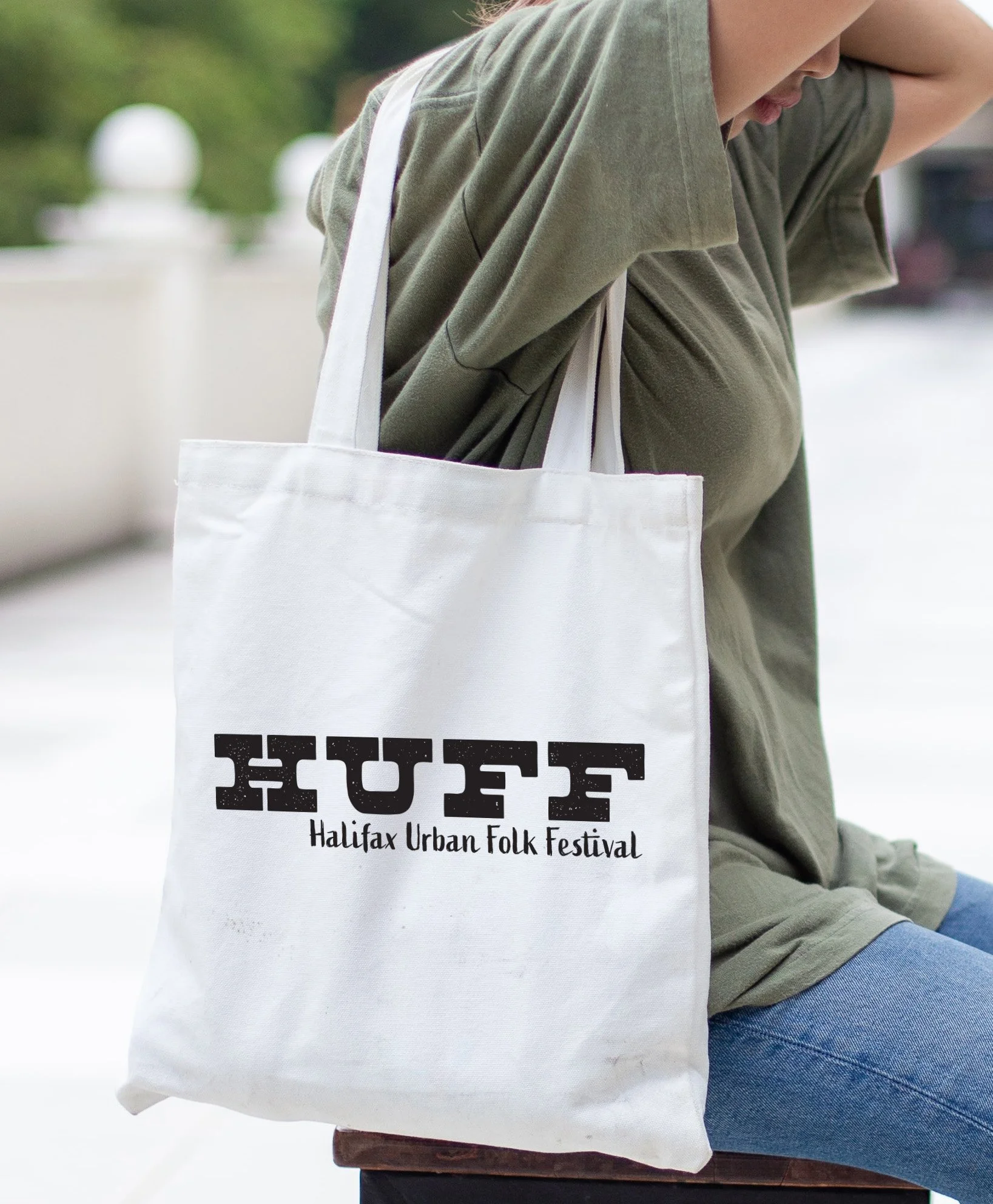 Person sitting outdoors holding a white tote bag with black text promoting the Halifax Urban Folk Festival.