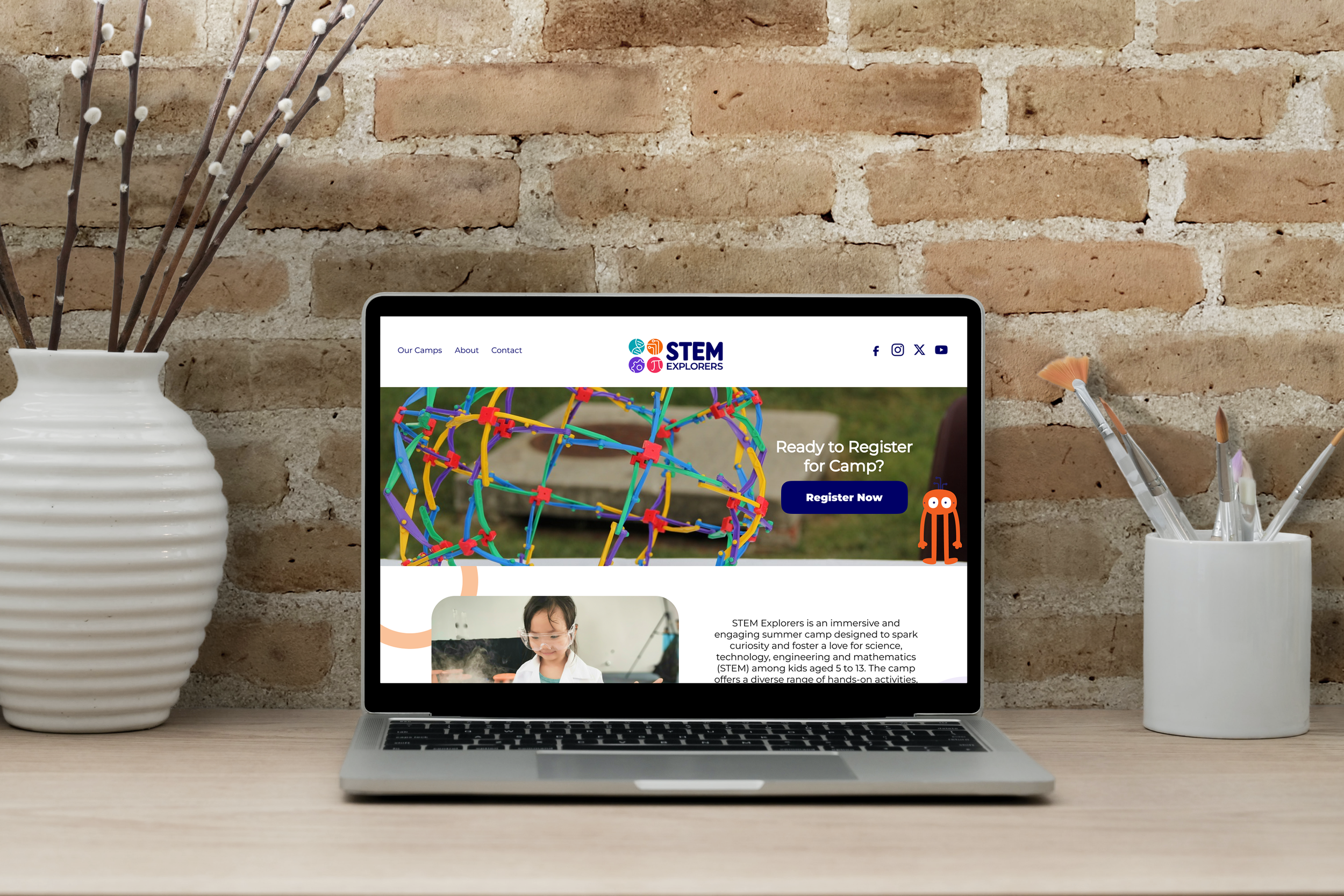 A laptop on a wooden desk displaying a STEM Explorers camp registration webpage, with a background of a brick wall and two white vases, one with brown twigs and the other with paintbrushes.