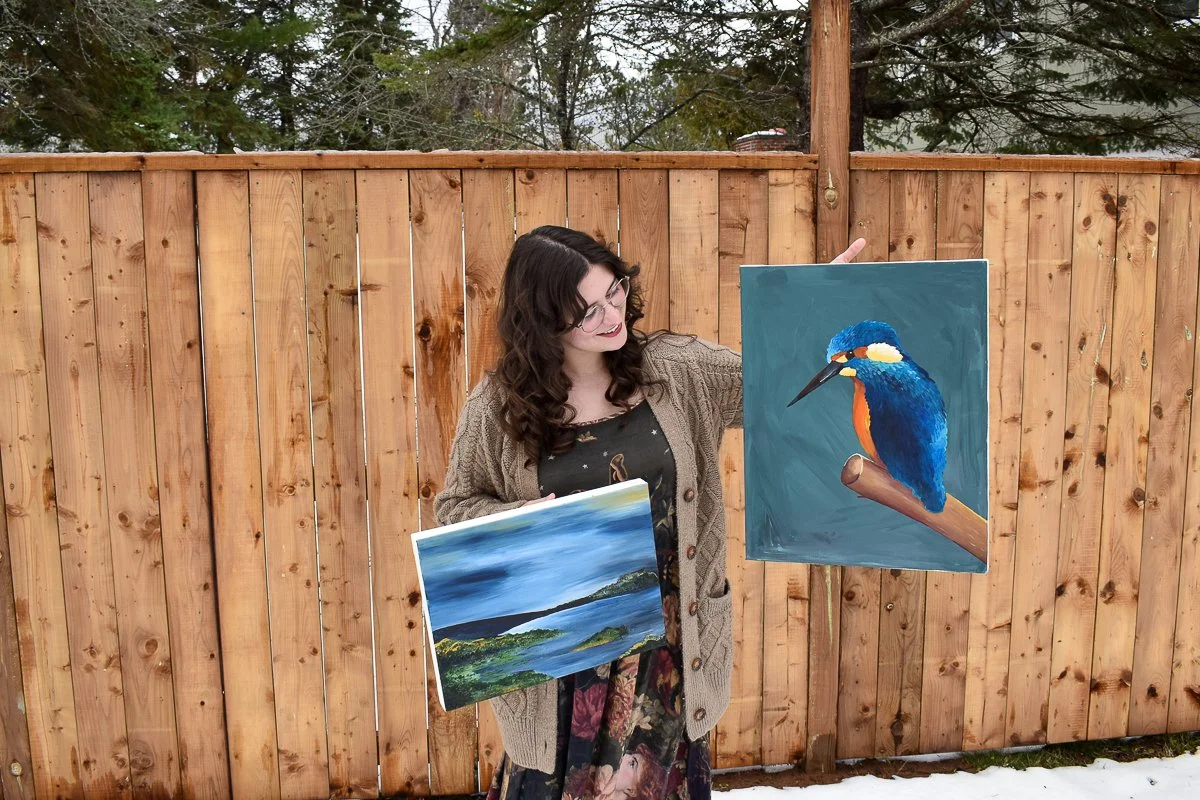 image of a women holding up two landscape paintings, standing in front of a fence.