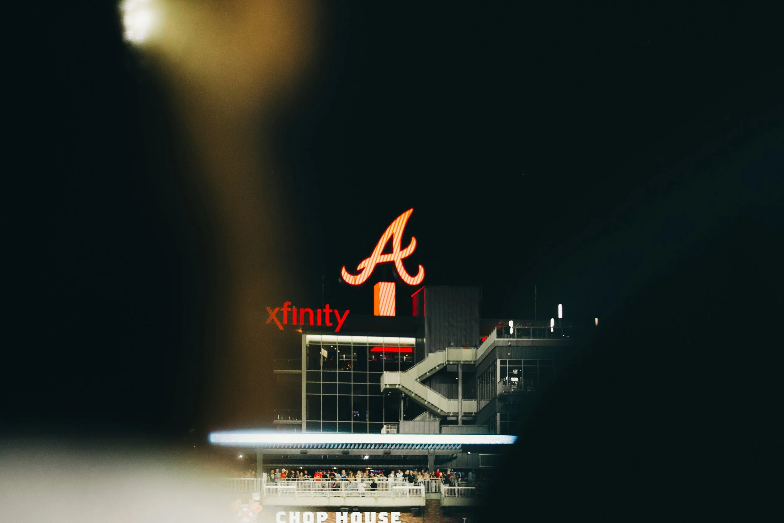 Nighttime view of a modern building with illuminated signage featuring the Atlanta Braves logo, an 'A', and the word 'xfinity' in red, with visible crowds on a terrace and a sign reading 'Chop House' at the bottom.