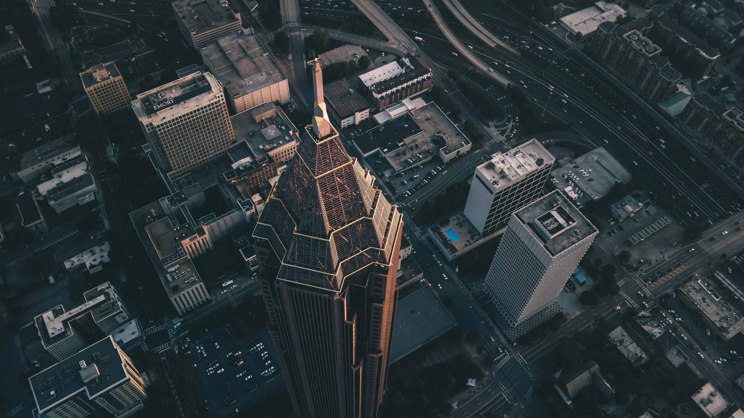 Aerial view of downtown Atlanta, Georgia, featuring a prominent skyscraper with a spire, surrounded by various office and residential buildings, with highways and roads visible.