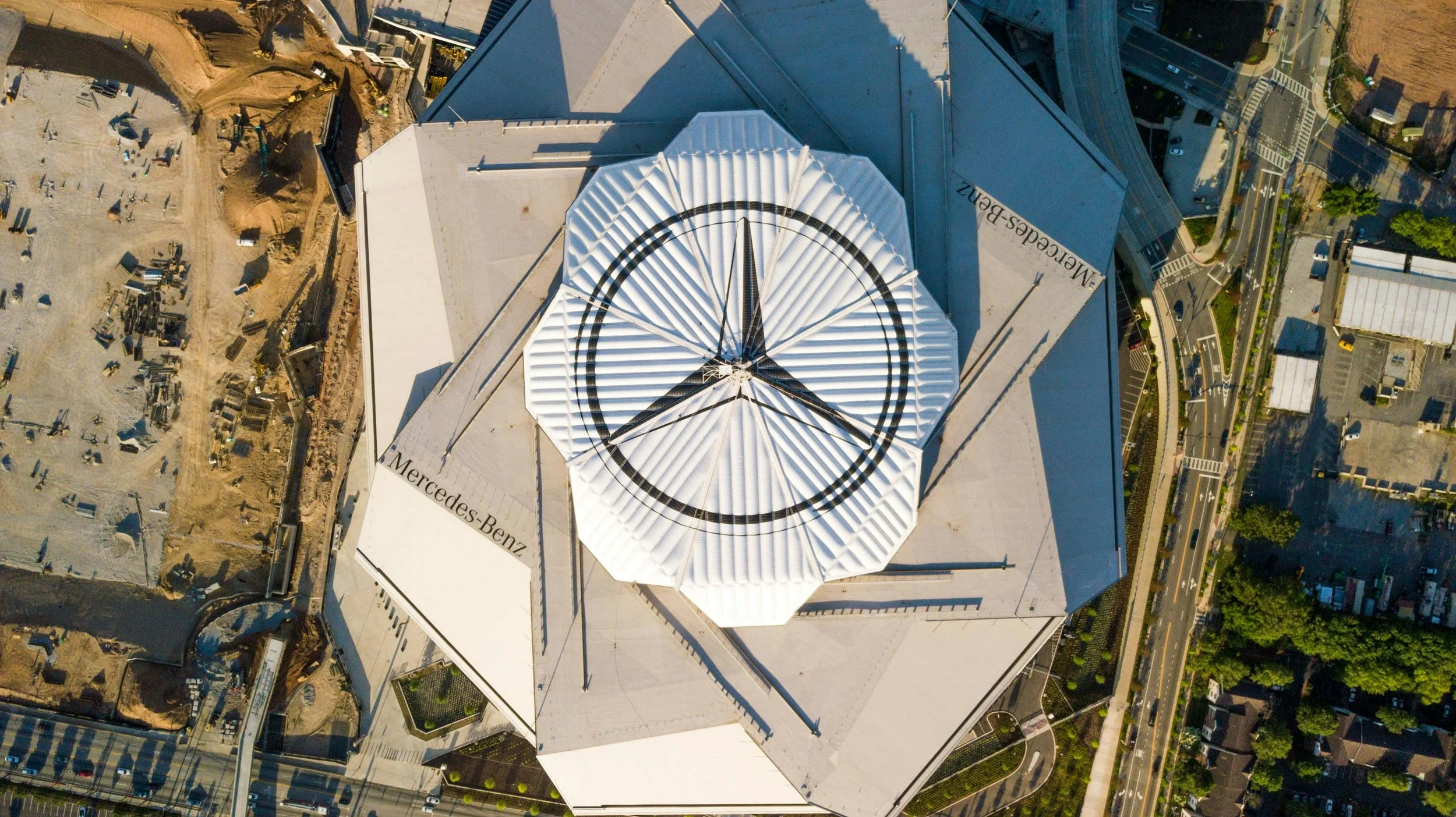 An aerial view of a building with a Mercedes-Benz logo on its roof, featuring a clock design, surrounded by roads, parking lots, and construction sites.
