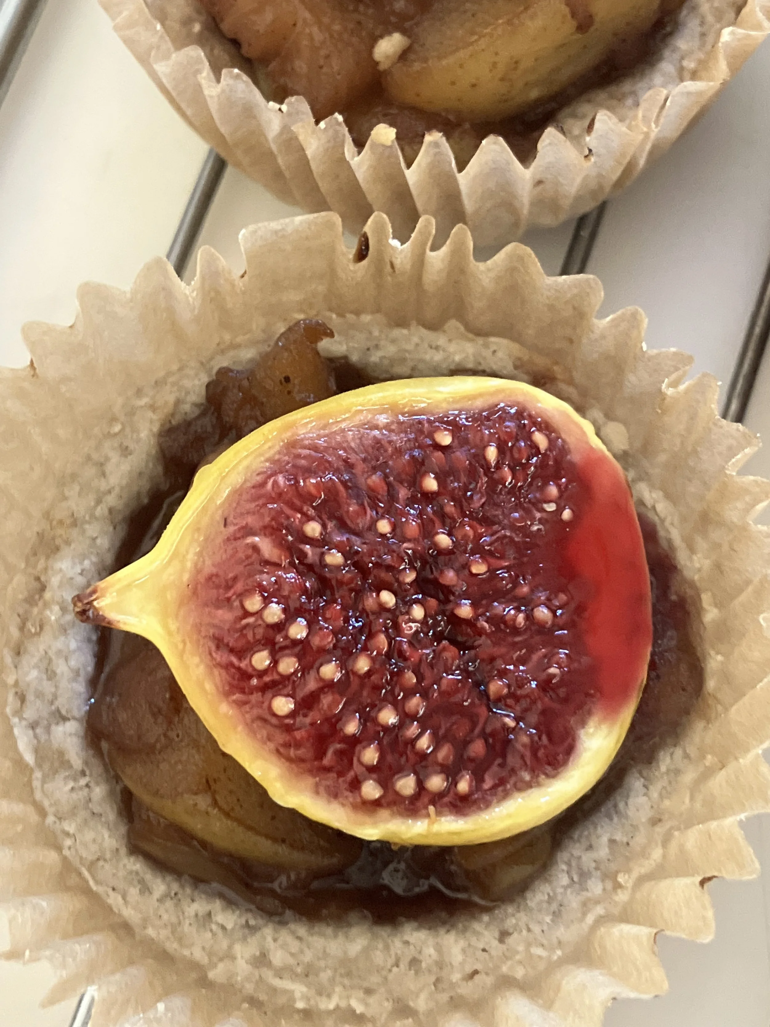 A fig half with a red interior and numerous seeds, placed on top of a tart or pastry in a paper cup, with other assorted baked goods in the background.