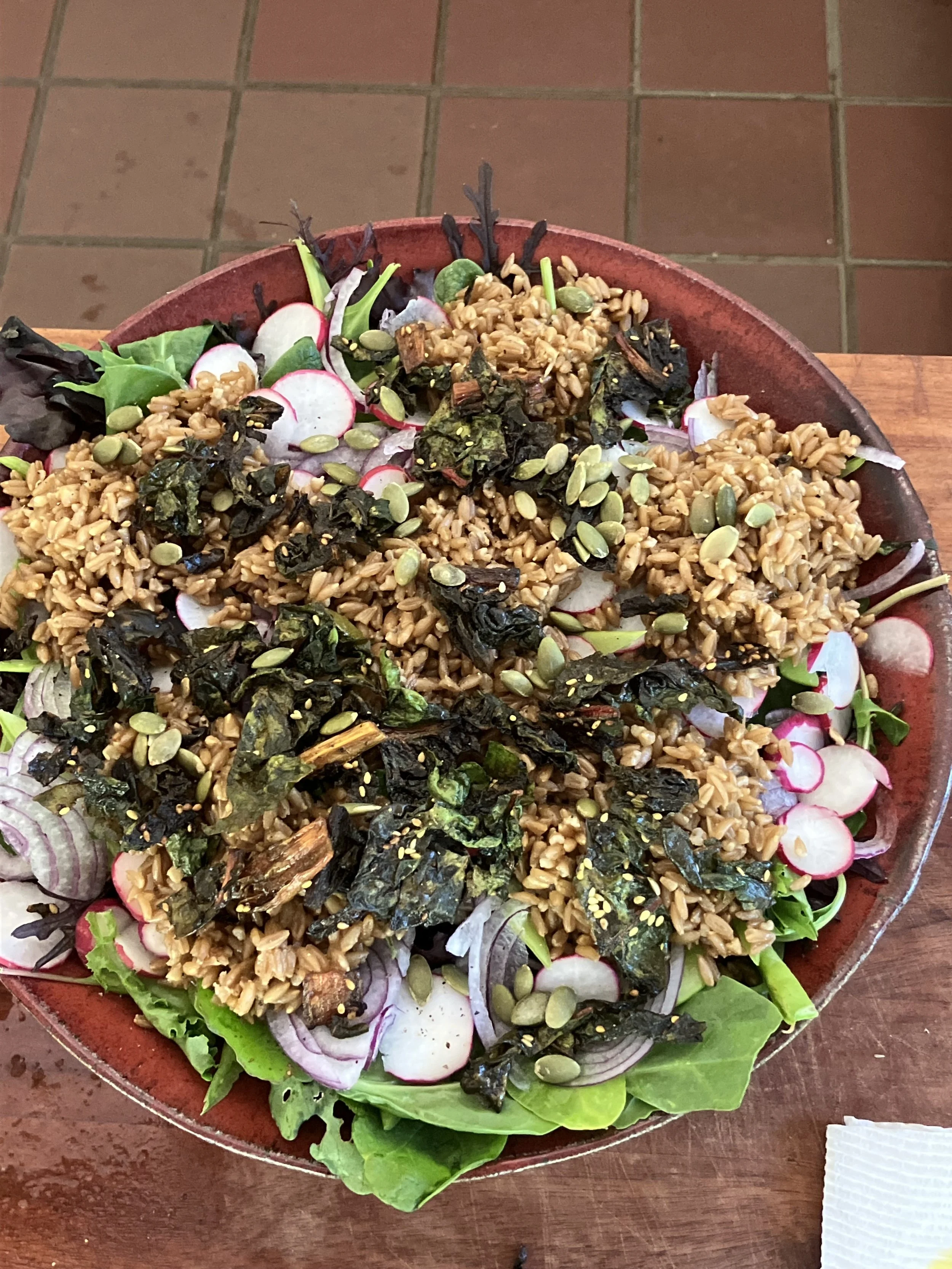 A large salad with mixed greens, radish slices, roasted rice, seaweed, and pumpkin seeds, served on a round wooden plate.