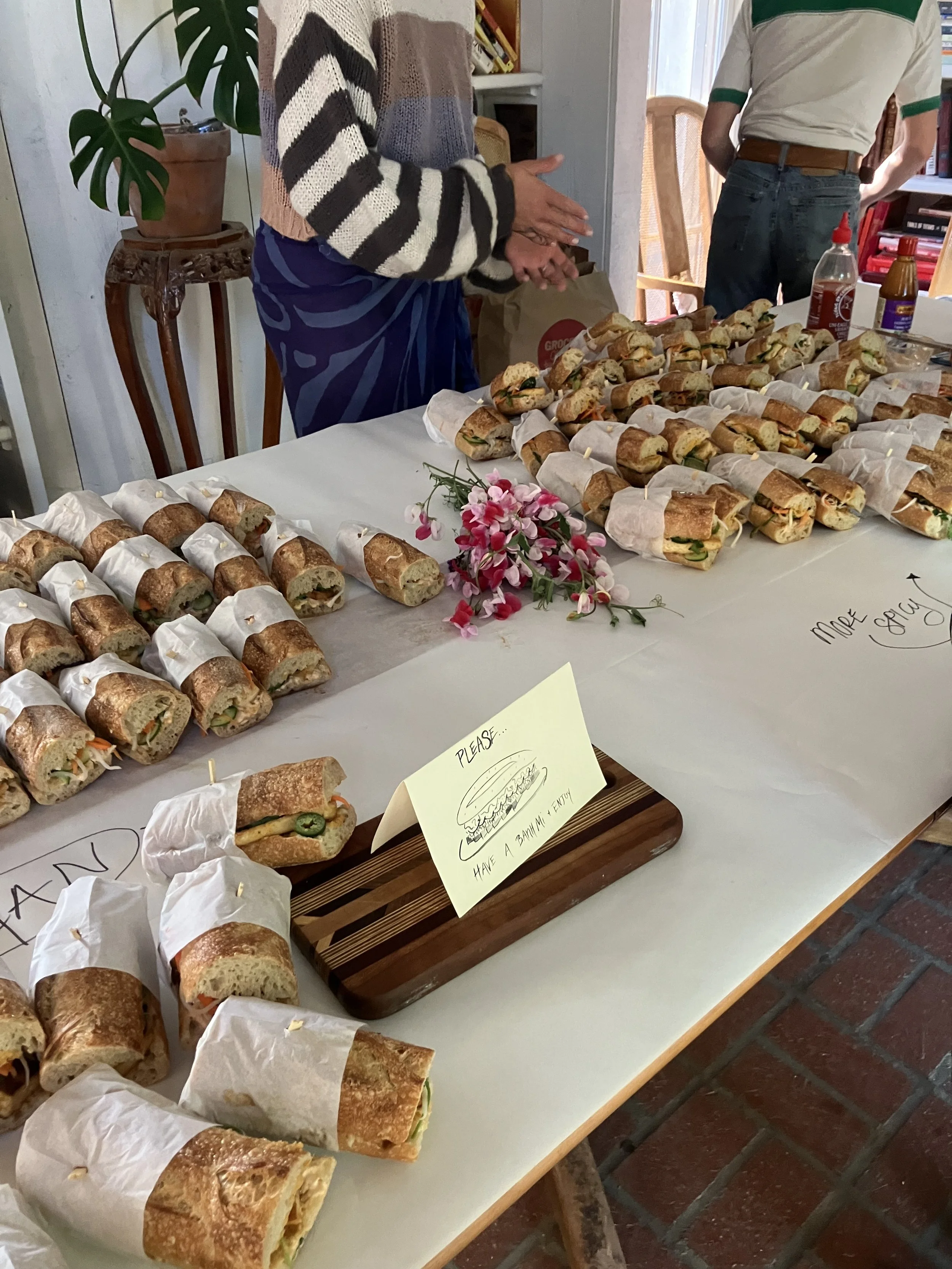 A table with numerous small sandwiches wrapped in paper and one unwrapped sandwich on a wooden tray with a sign that reads 'Please Have a Smile & Enjoy.' Two people are standing behind the table, one gesturing with hands and the other with arms crossed, with a bookshelf and window in the background.