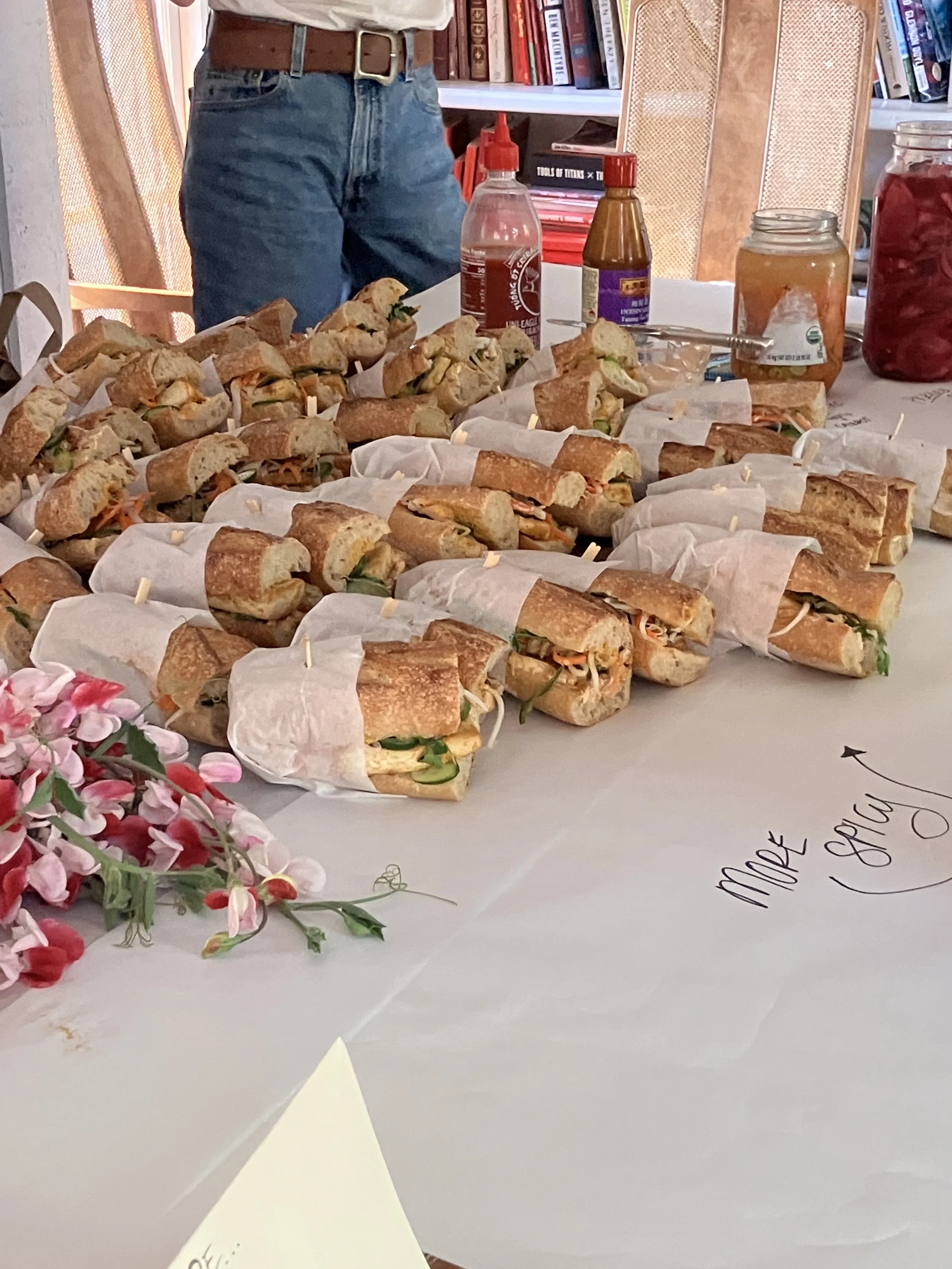 Table filled with assorted sandwiches wrapped in paper, with a bouquet of pink flowers on the side. In the background, there are bottles of condiments, jars, and books on a shelf.