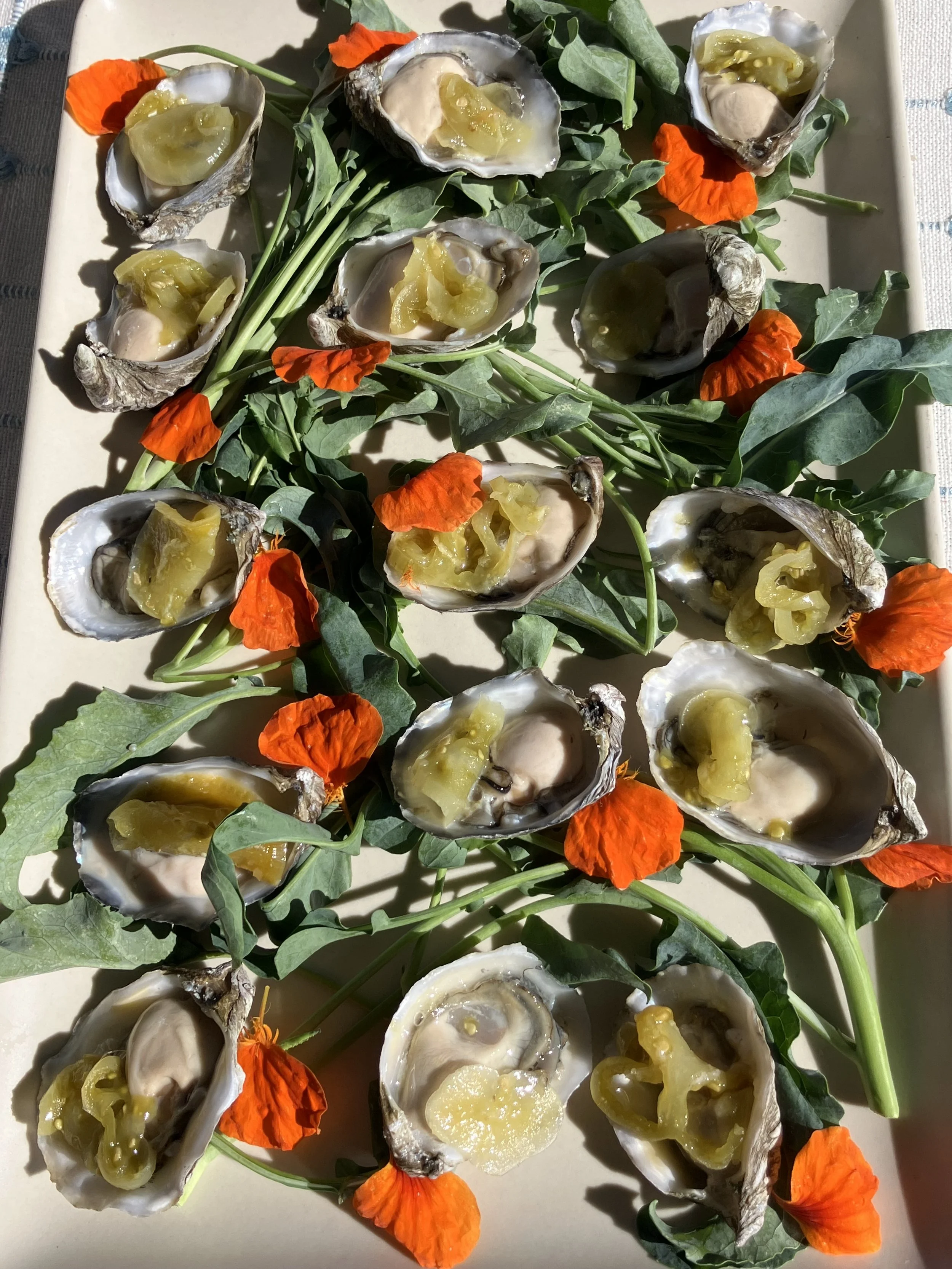 Oysters served on a platter garnished with green leaves and orange edible flowers, with lemon or similar citrus fruit in each oyster.