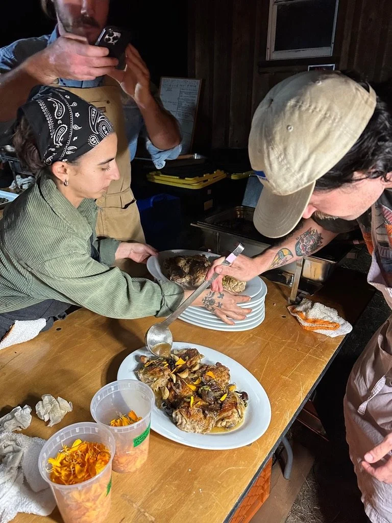 People serving and preparing a roasted meat dish on a kitchen table with garnishes.