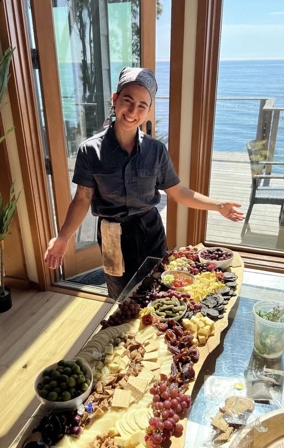 Smiling person in dark gray chef's outfit and head covering standing next to a long table with a large charcuterie and cheese board, with a background of sliding glass doors and a view of the ocean.