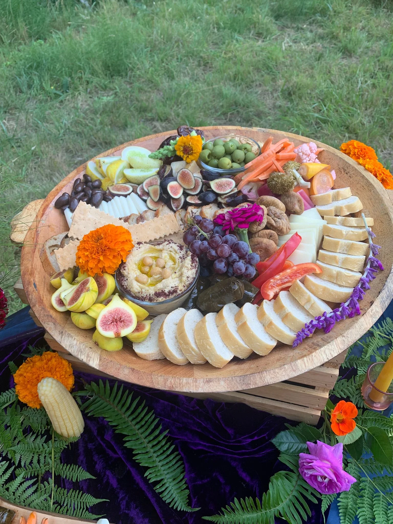 A round wooden cheese and fruit platter with various cheeses, grapes, figs, nuts, carrots, and decorative flowers, set on a purple velvet cloth outdoors.