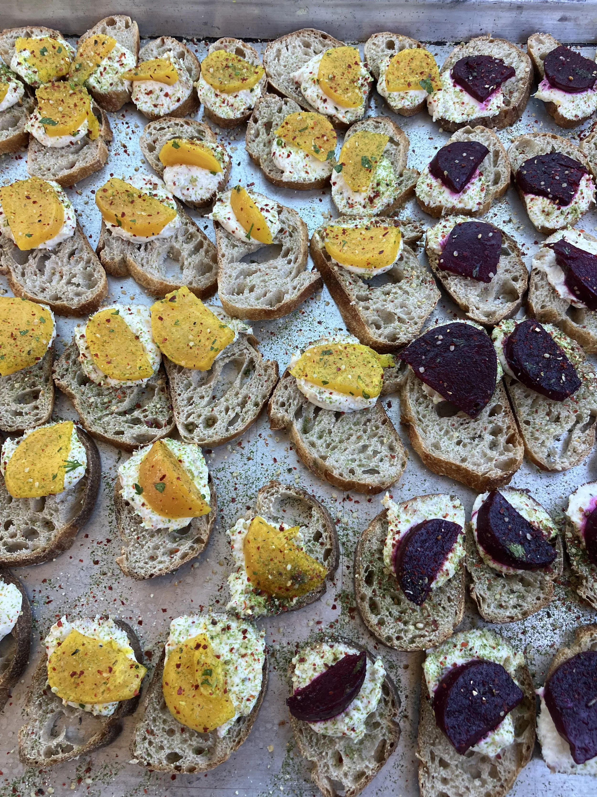 Assorted slices of baguette with cream cheese, topped with pickled fruit and sprinkled with spices, arranged on a tray.