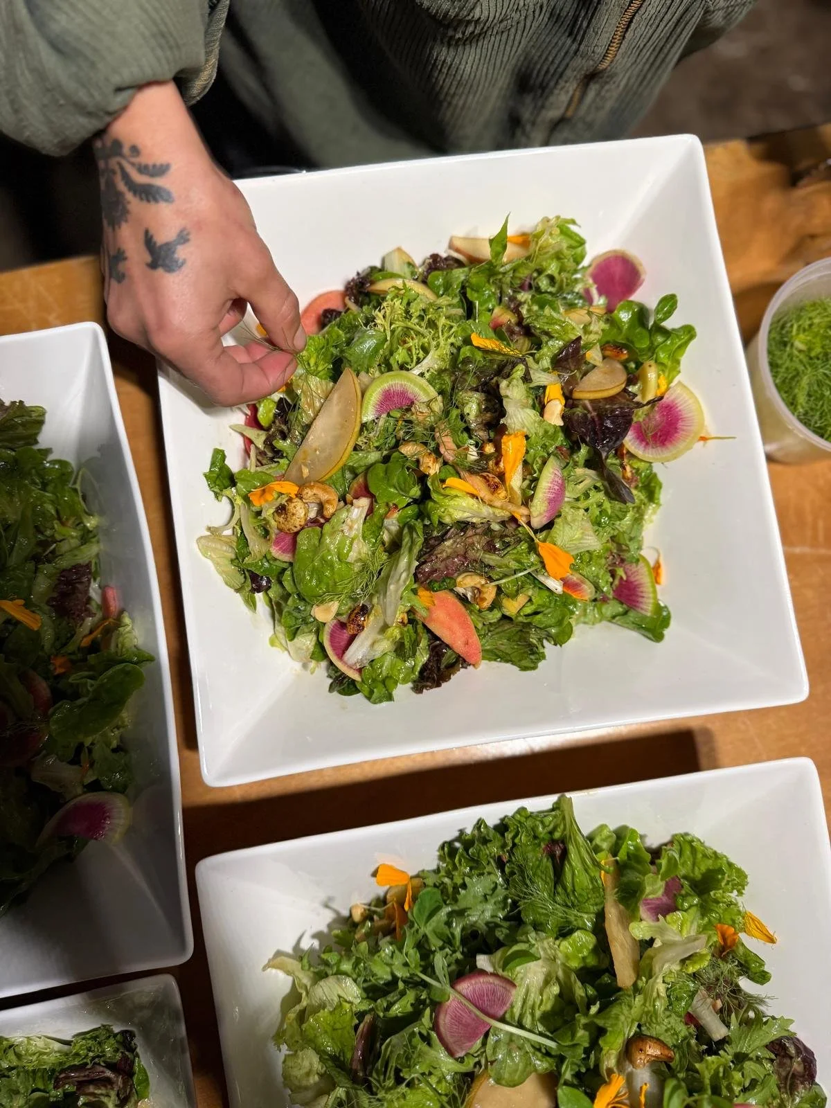 A large square white bowl filled with a fresh mixed green salad containing watermelon radishes, edible flowers, and nuts, with a person's hand garnishing the salad. There is also a smaller bowl of green salad and a cup of what appears to be herbs or sprouts on a wooden table.
