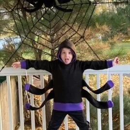 Child with four spider legs costume standing on porch with railing and spider web decoration in the background