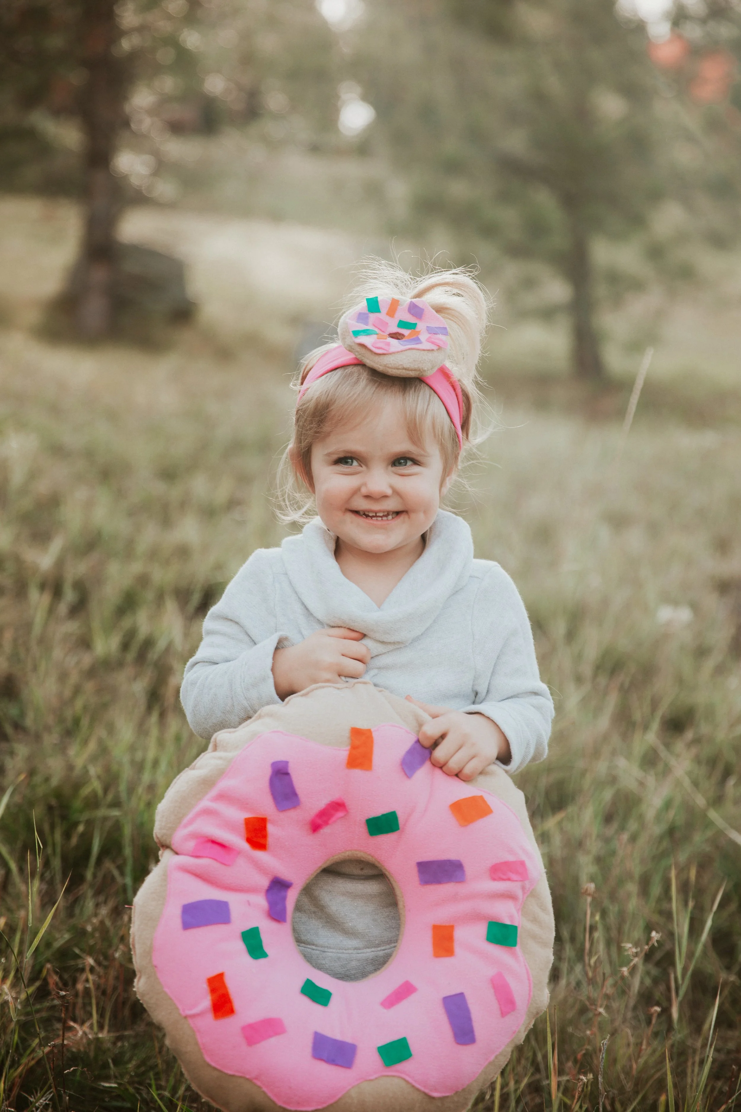 A young girl smiling outdoors while holding a large plush donut with pink frosting and colorful sprinkles. She is wearing a gray hoodie and a pink headband with a matching plush donut on top.