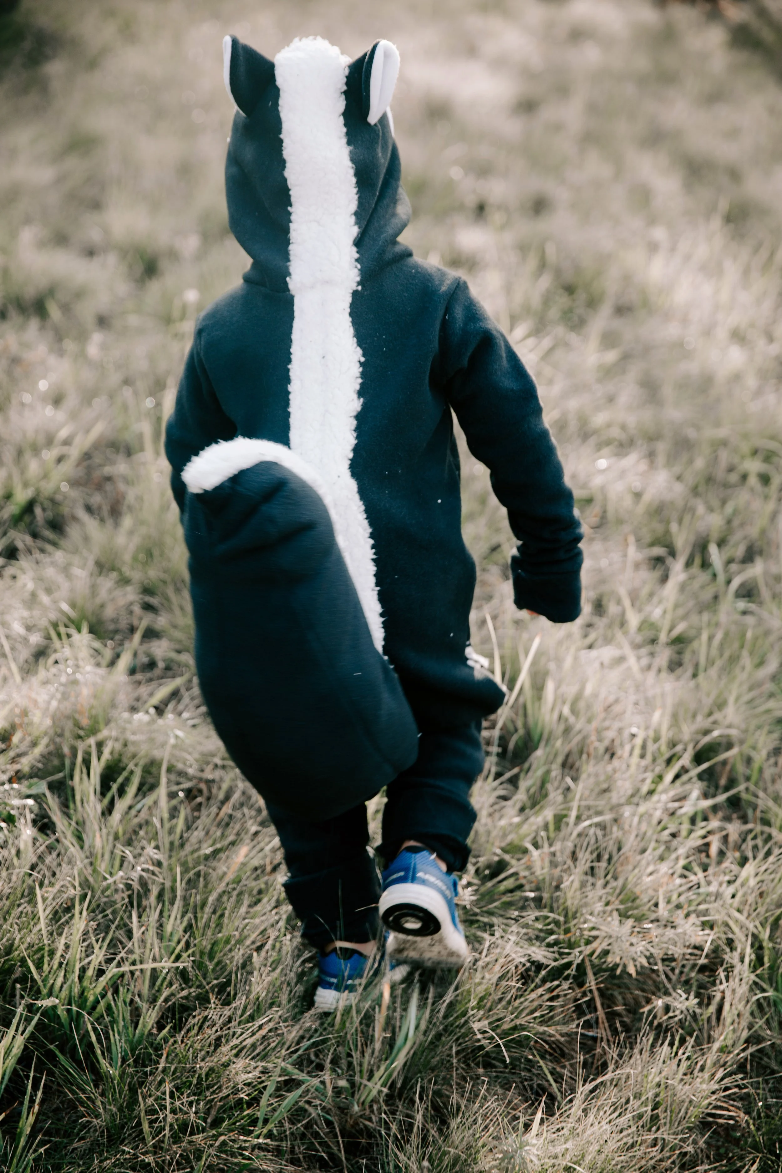 Child in a black hoodie with a sheep costume hood, running through a grassy field with blue sneakers.