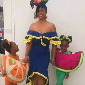 Woman and two children dressed in fruit-themed costumes, smiling in front of a white wall.