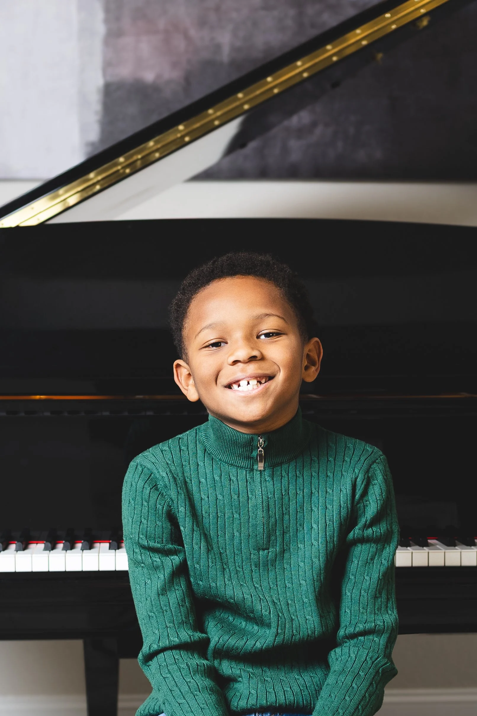 A smiling young boy wearing a green sweater sitting in front of a black piano.