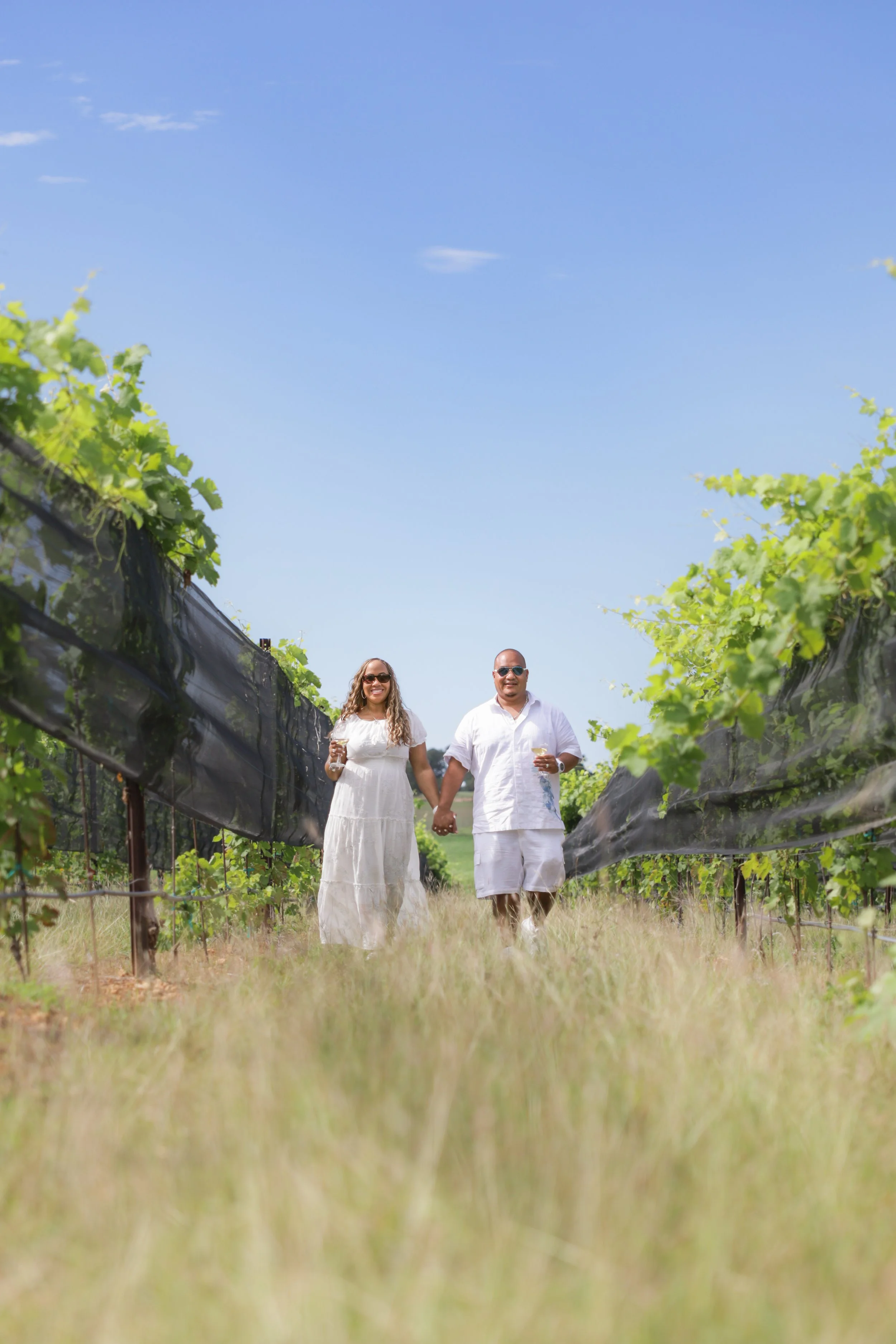 A couple walking hand in hand through a vineyard on a sunny day, both holding glasses of wine.