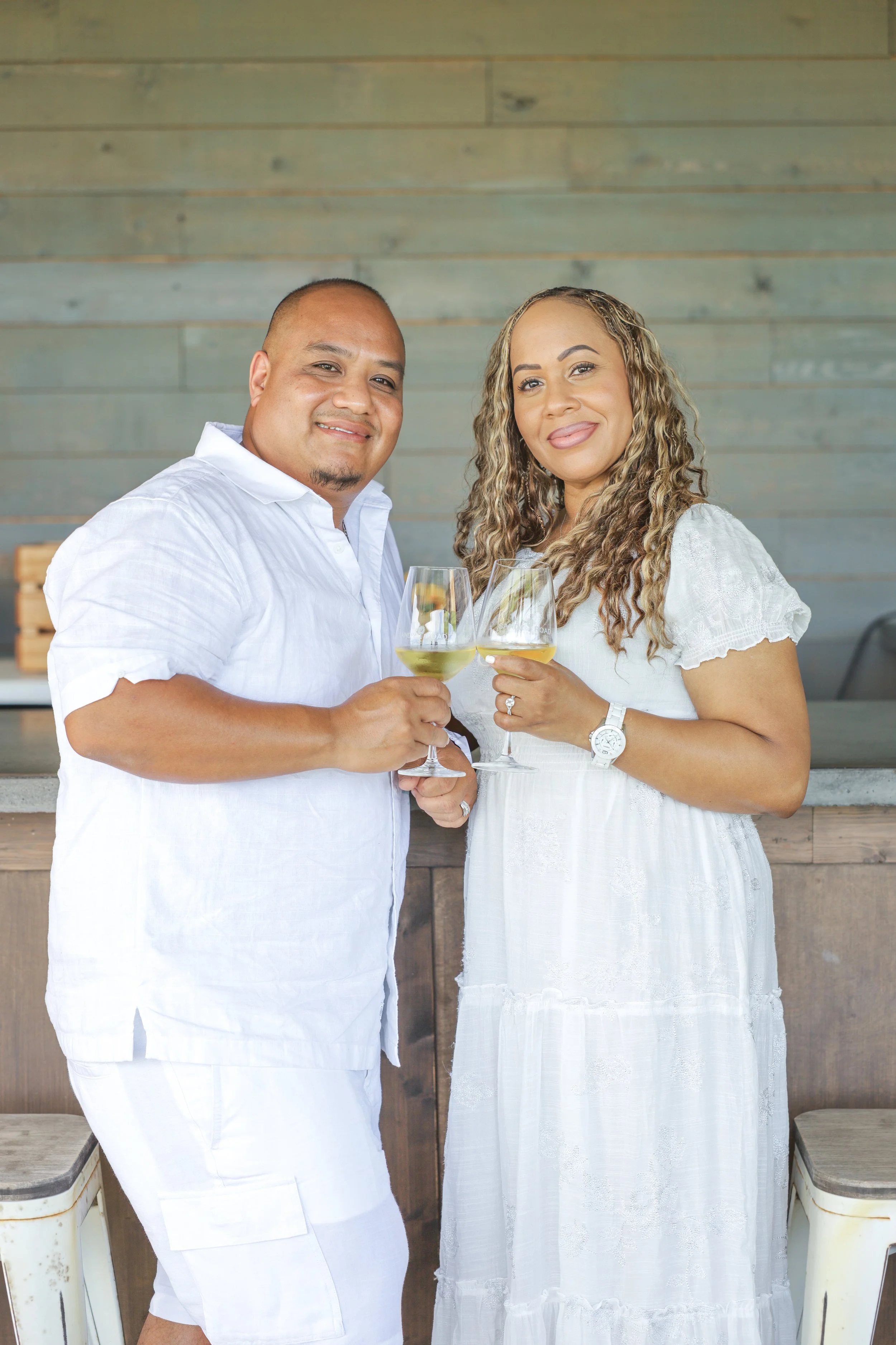 A couple dressed in white, smiling and holding glasses of white wine, standing outdoors against a wooden wall.