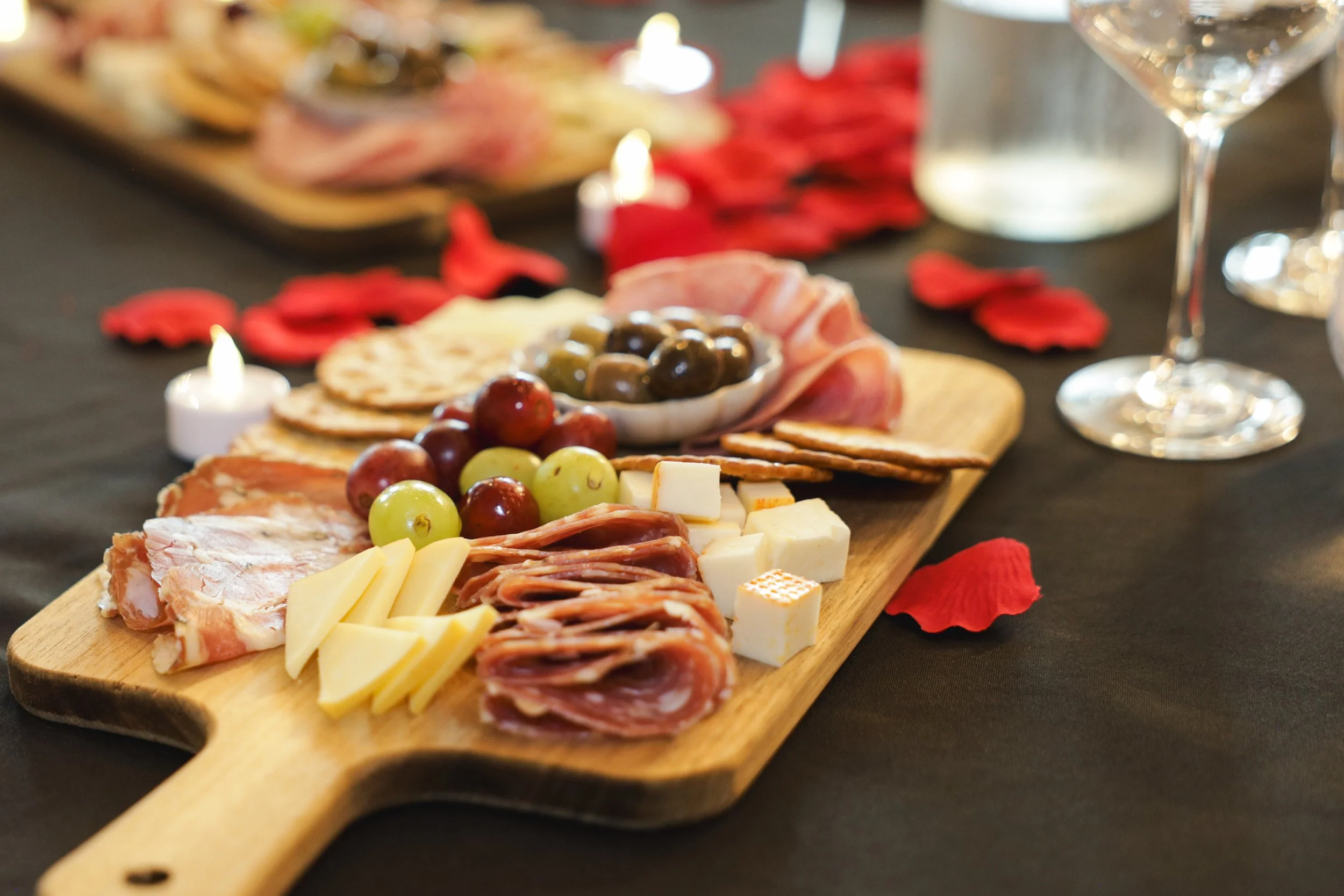 A wooden serving board with assorted cheeses, grapes, sliced meats, and a bowl of green and black olives on a black tablecloth.
