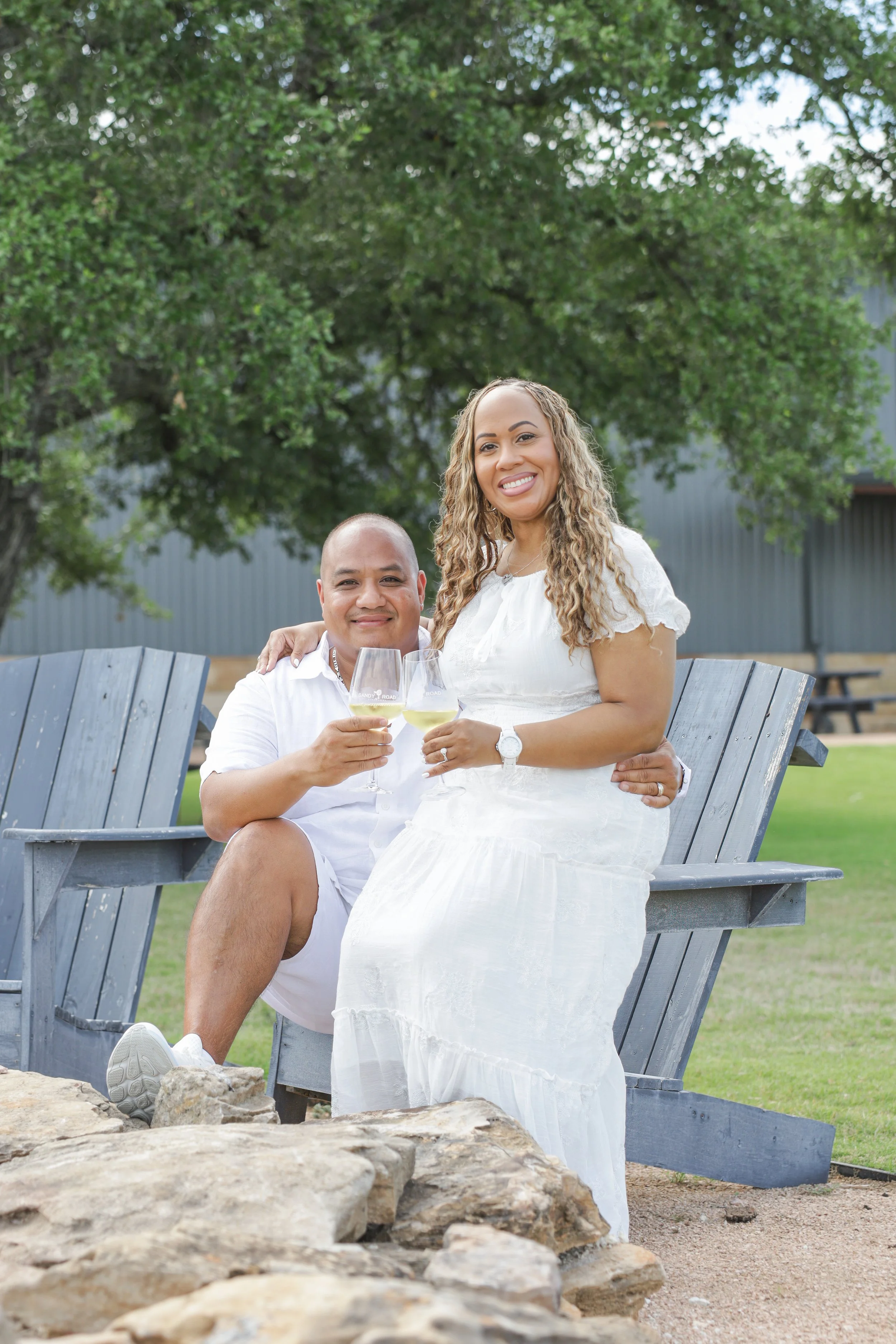 A smiling woman in a white dress and a smiling man in a white shirt and shorts sit on a bench outdoors, holding wine glasses. They are surrounded by green trees and rocks in the foreground.